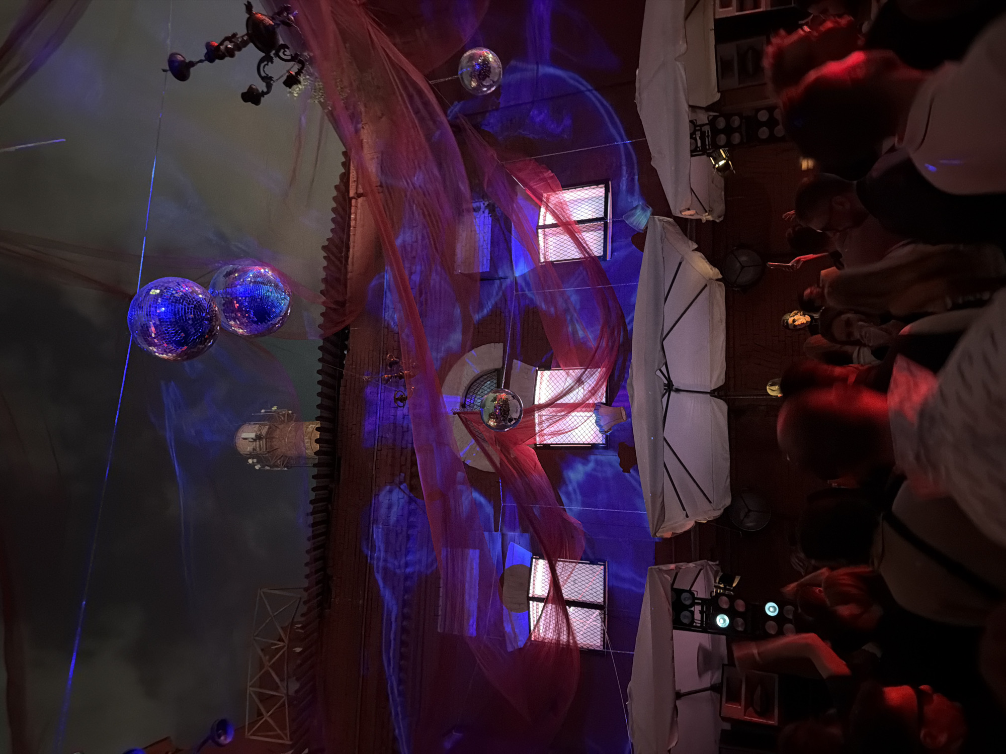 The photograph shows the interior of a building in Moscow, Russia. Two mirrored disco balls hang from the ceiling on the left. A complex chandelier is also visible in the upper left. The walls are made of brown brick and feature rectangular windows with metal grates. Sheer red fabric is draped across the walls. A single mirrored disco ball hangs centrally in the middle of the image. Blue lighting effects are projected onto the wall. On the right, a vertical row of white parasols is positioned along the wall. People are visible on the far right of the photograph, though they are obscured. The angle of the photograph suggests it was taken looking upwards, as if from the floor of the building.