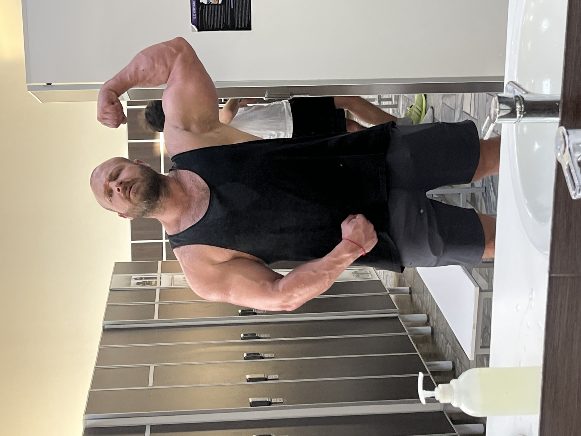 A bald, bearded man in a black tank top and grey shorts poses in a gym locker room, flexing his right bicep. He is standing in front of a row of lockers. A white sink and a soap dispenser are visible in the foreground, reflected in a mirror. Another man in a white t-shirt and black shorts can be seen in the background, also reflected in the mirror. The floor of the locker room is tiled. The image is taken at the "Fitness" fitness club in Chaikovsky, Russia.