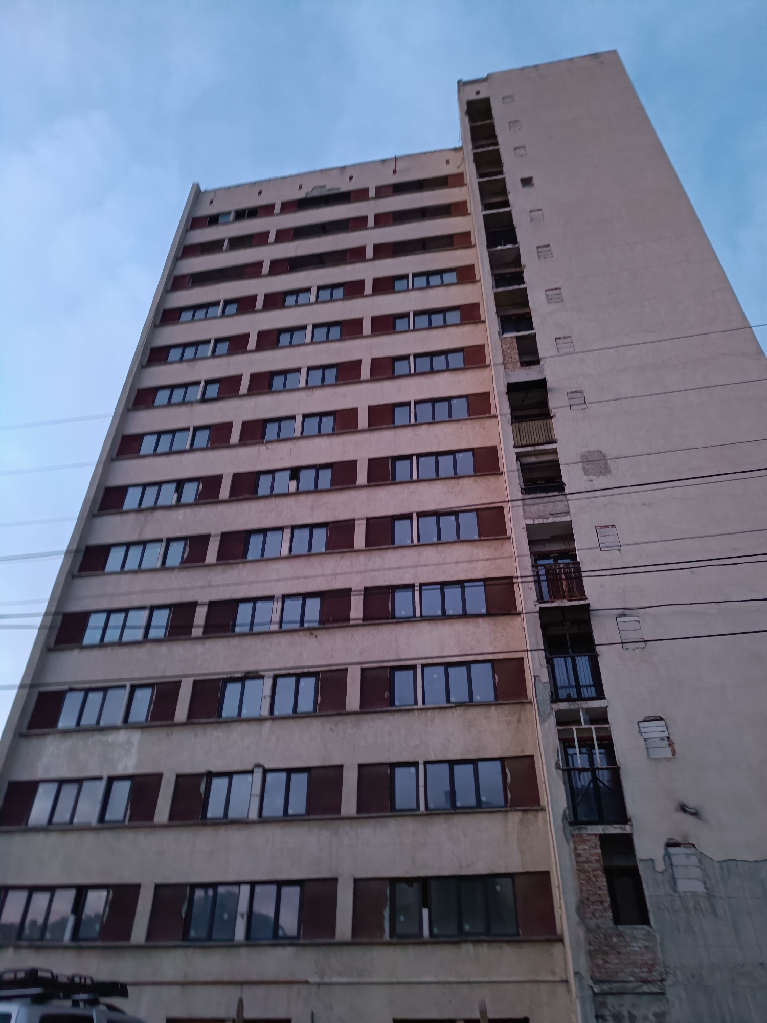 The image displays a tall, multi-story apartment building, seen from a low-angle perspective looking upwards. The building dominates the frame, set against a soft, light blue sky that shows subtle hints of pink or orange, suggesting either dawn or dusk.
The primary facade of the building is light beige or off-white, characterized by a regular grid of rectangular windows. These windows mostly have dark frames and reflective glass, beneath which are dark brown or reddish-brown horizontal panels. The building's left side features this consistent architectural pattern, with its edge tapering slightly as it ascends.
In stark contrast, the right side of the building appears to be either undergoing significant renovation or is in a state of disrepair, or perhaps even partial demolition. This narrower section features a series of vertical cutouts or former balconies, some of which are now empty voids with exposed structural elements, while others show patched-up brickwork or remnants of railings and window frames. Patches of missing plaster and exposed concrete are evident on this side. Multiple dark utility or power lines stretch horizontally across the middle and lower parts of this damaged facade.
No people or explicit activities are visible, lending a static quality to the scene. The architecture, including the uniform facade and the signs of wear or renovation, is consistent with urban buildings found in Pleven, Bulgaria, as indicated by the context. A small portion of a vehicle's roof rack is partially visible at the bottom left edge of the frame. No text is visible in the image.