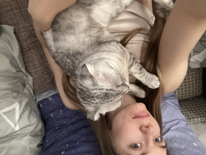 The scene, located in Zvezda, Russia, depicts a young adult female lying down, holding a domestic cat. The female, possessing long brown hair and fair skin, wears a light-colored tank top and gazes directly at the camera with a neutral expression. A light gray tabby cat with distinct darker stripes rests horizontally on her upper torso and left shoulder, with its head turned away from the camera and appearing to be asleep or resting. The immediate background includes a dark blue pillow with white speckles behind the female's head and a light gray duvet with a prominent white triangular pattern on the right side of the frame. A portion of a light-colored woven basket is visible on the far left. The subjects are situated on a textured, brown fabric surface, indicative of a bed or sofa in an indoor environment.