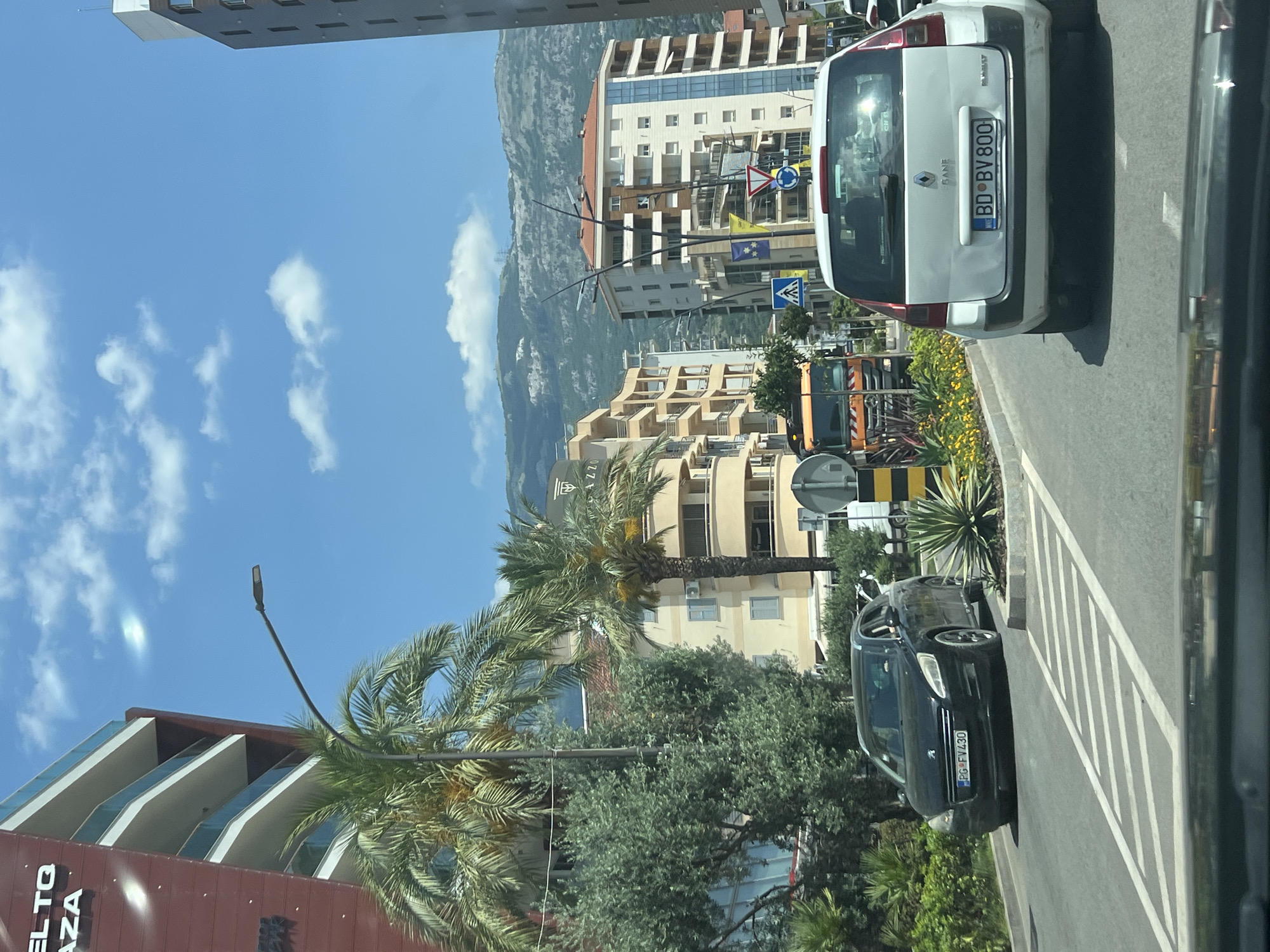 The image depicts a street scene in Budva, Montenegro. A white Renault station wagon with license plate "BD BV 800" is in the foreground, partially obscuring a black Peugeot sedan with license plate "PG FV430." Both vehicles are on a paved road with white lane markings.
To the left of the road, a modern building with a red facade and balconies is visible, along with palm trees and lush greenery. Further down the street, a yellow and orange construction truck is stopped. In the background, multi-story apartment buildings with light-colored facades line the street, and a mountain range stretches across the horizon under a bright blue sky with scattered clouds. A streetlamp is visible on the left side of the road. The scene appears to be a sunny day in an urban environment.