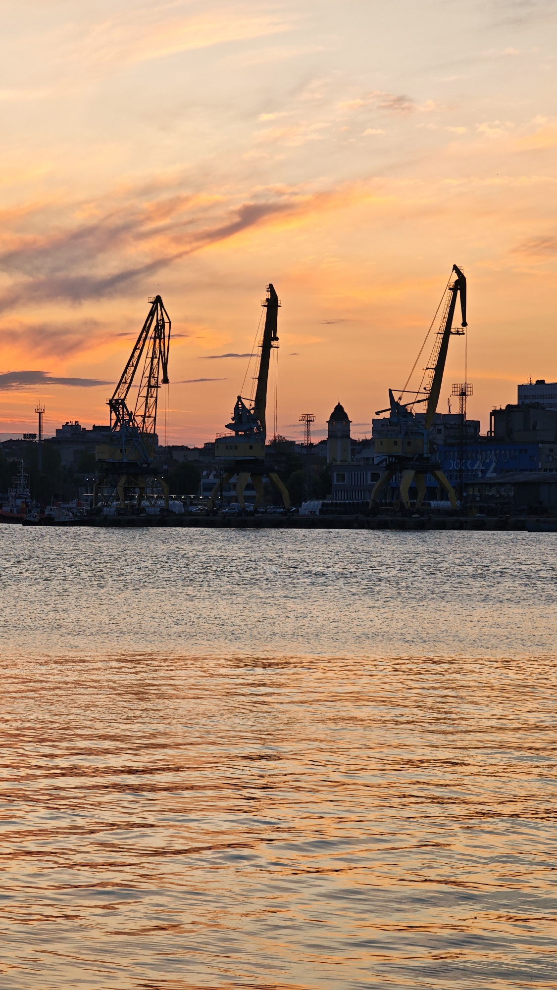 The image depicts a waterfront scene, likely a harbor or port area, at sunset or sunrise. The sky is filled with shades of orange, red, and pink, indicating a time of day when the sun is near the horizon. Silhouetted against the bright sky are several cranes, characteristic of a port facility. These cranes dominate the middle ground of the image. Behind the cranes, there is a suggestion of a city skyline, with buildings and structures faintly visible. The foreground is dominated by water, reflecting the colors of the sky. The water has small ripples or waves.
Given the prompt specifying the location as Burgas, Bulgaria, it's probable that this is the Port of Burgas.