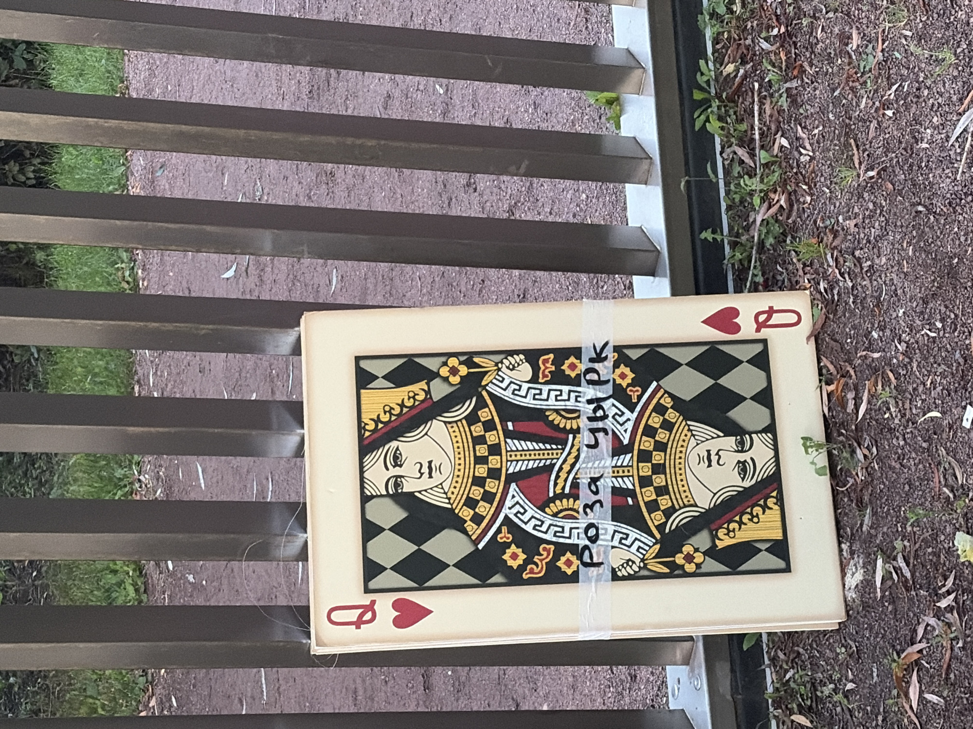 An art installation, a large playing card depicting the Queen of Hearts, leans against a metal fence. The card is positioned upright, leaning back slightly. On the card, a handwritten label reads "Роза Чыpk". The fence is made of vertical metal bars, some of which are partially obscured by the card. The ground in front of the fence is covered with dirt, leaves, and sparse green vegetation. In the background, grass and some foliage are visible. The scene appears to be in an outdoor public space, possibly a park or walkway. The lighting suggests it is daytime.