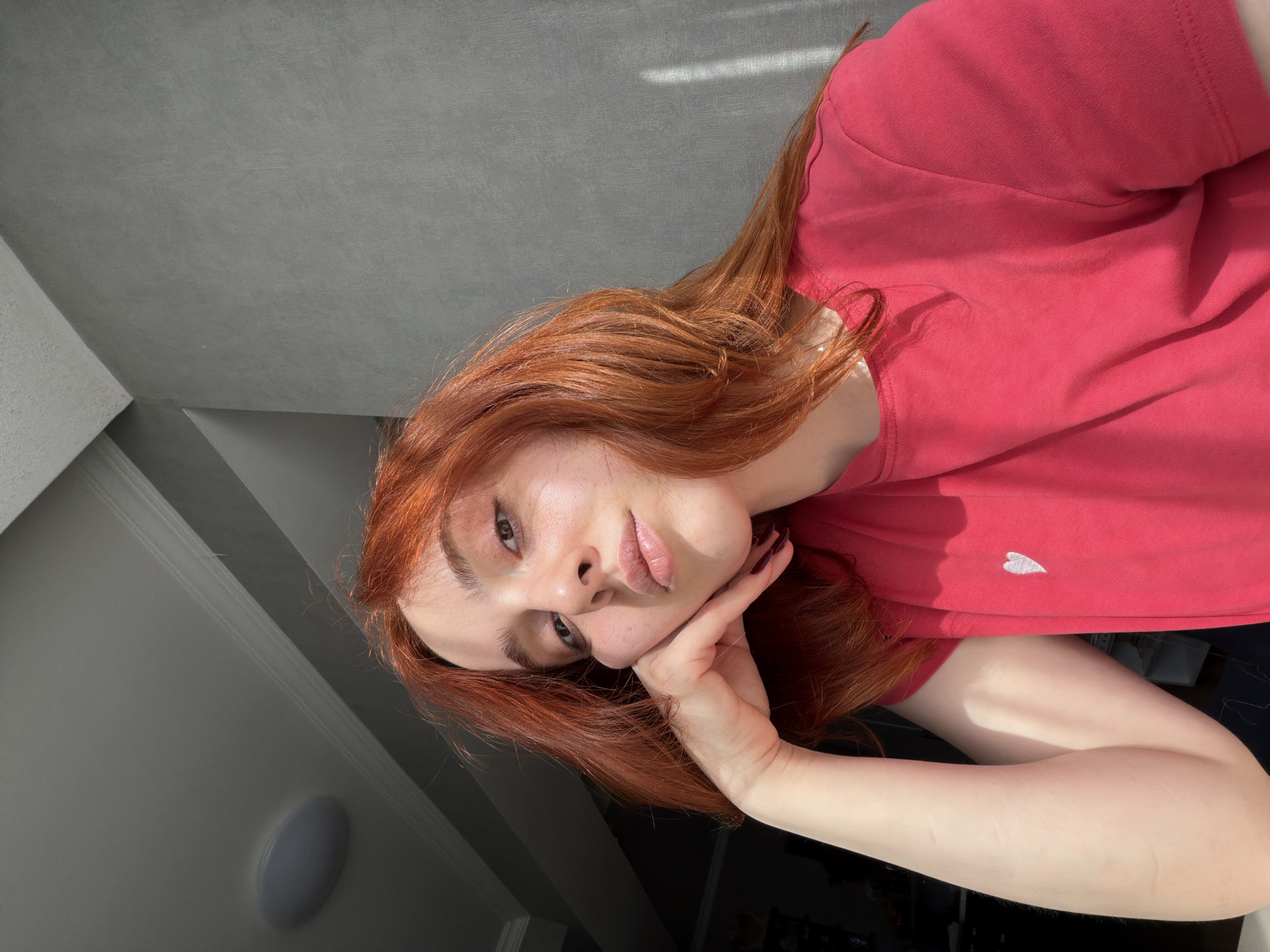 A young woman with red hair is resting her head on her hand in what appears to be an indoor setting, possibly an apartment or house. The woman is fair-skinned, and she's wearing a pink shirt with a small white heart detail. She has light makeup, emphasizing her lips. Her red hair falls around her face and down her arm. The background includes a gray ceiling and white walls, suggesting a modern interior. The overall lighting suggests a sunny day. There are no specific details indicating that the location is in Tbilisi, Georgia.