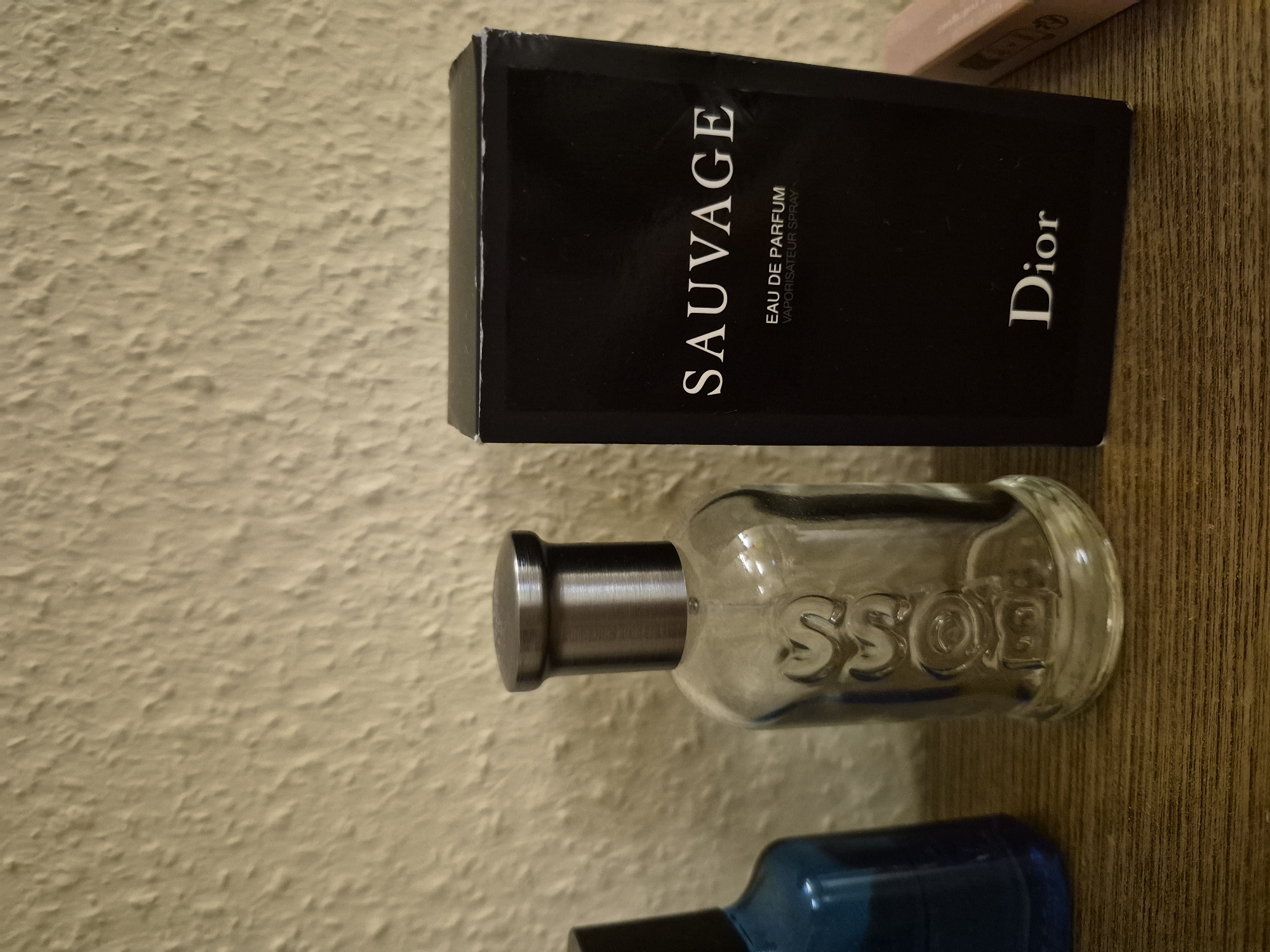 A perfume bottle with a metal cap and the embossed word "BOSS" on the glass is placed next to a black box with "SAUVAGE EAU DE PARFUM VAPORISATEUR SPRAY" and "Dior" printed on it. The items are on a wooden surface against a textured, light-colored wall.