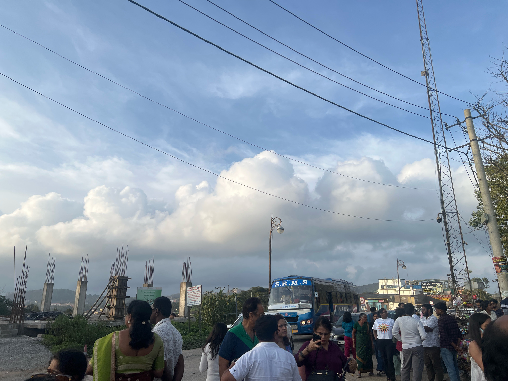 This image captures a street scene in Madikeri, India, likely during daylight given the visible sky and lighting. In the foreground, a crowd of people is gathered, suggesting a public area or a bus stop. Several individuals are looking in different directions, engaged in conversation or observing their surroundings. A blue and white bus with "S.R.M.S" printed on it is visible in the mid-ground, indicating a transportation hub or a route passing through this location.
Behind the crowd, construction is underway, evidenced by concrete pillars and rebar rising from a foundation, suggesting ongoing development or infrastructure work. Overhead, power lines crisscross the frame, supported by utility poles and a taller telecommunications tower on the right. The sky is a mix of blue and dramatic, billowing white clouds. In the background, buildings and some greenery are discernible, hinting at the urban or semi-urban character of Madikeri. The overall impression is one of a busy, developing area with elements of everyday life, infrastructure, and construction.