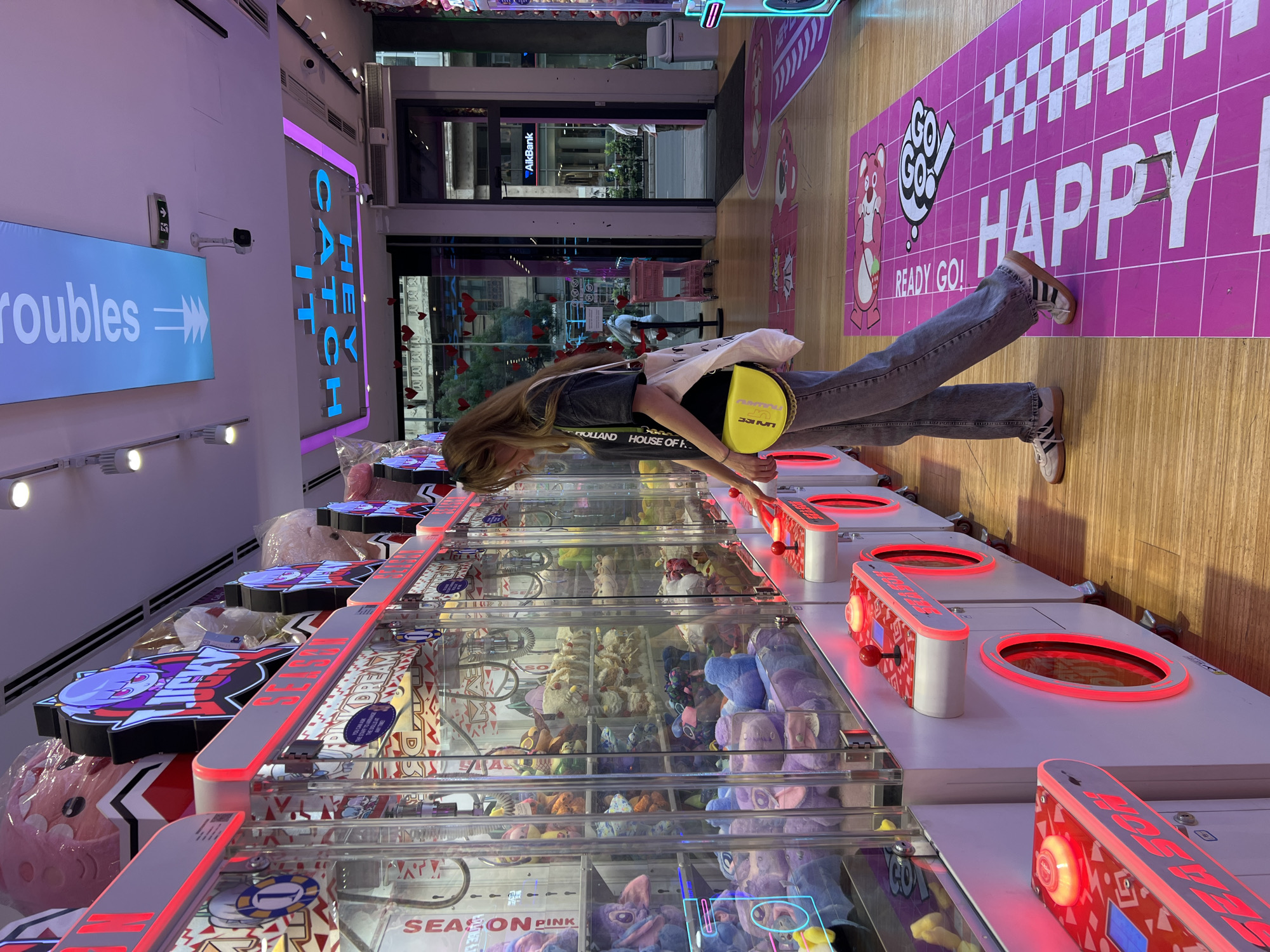 A young person with long blonde hair stands at a row of claw crane arcade machines in a brightly lit indoor arcade, potentially located in Belgrade, Serbia. The machines are filled with various plush toys. The person is wearing a denim shirt, jeans, and sneakers, and has a yellow and white bag slung over their shoulder. On the left, a sign reads "Hey Catch Troubles," and a pink floor graphic says "Happy." Through the arcade's glass doors, a street scene is visible, showing an "AikBank" sign, as well as building facades and red heart decorations. The overall atmosphere is colorful and playful.