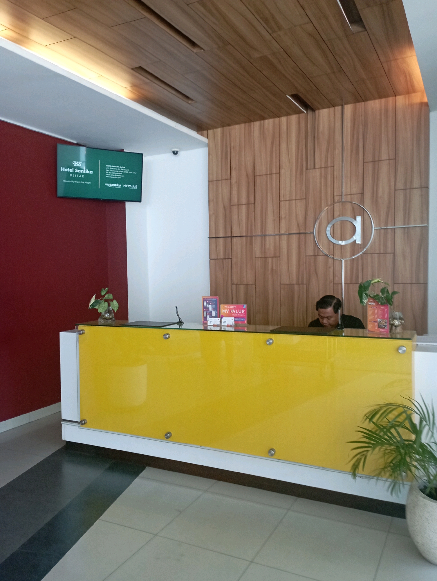 The image shows the reception area of the Hotel Santika Blitar, located in Malang, Indonesia. A male receptionist, wearing a dark shirt, is seated behind a bright yellow reception desk, looking down at a laptop. The desk is topped with a glass surface, on which several items are placed, including two small potted plants, a black folder, and small display stands with promotional material. Behind the desk, a wall is decorated with vertical wooden panels and a circular logo featuring the letter 'a'. To the left of the reception desk, a large screen displays information about the Hotel Santika Blitar, including its address and contact details, along with the "mysantika MYVALUE" logo. The surrounding environment features a maroon-colored wall on the left, white walls, a wood-paneled ceiling with recessed lighting, and a tiled floor with a contrasting pattern. A potted palm tree stands to the right of the reception desk.