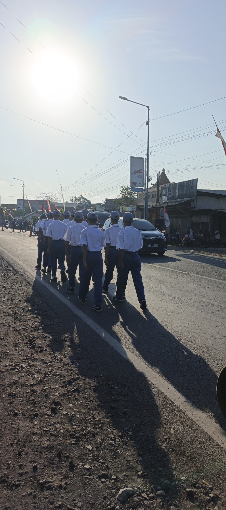 The image depicts a group of Indonesian students in school uniforms marching along a paved road in Kasiyan, Indonesia. The students, all wearing white short-sleeved shirts, dark blue trousers, and grey caps, are walking in a single file line away from the camera. Their shadows stretch long across the asphalt, indicating a low sun angle, likely during the early morning or late afternoon.
The road appears to be a two-lane street in a semi-urban or rural area, with a rough, gravelly shoulder on the left side of the image. In the background, there are buildings lining the right side of the road, including a structure with a sign that appears to read "POM BENSIN" (gas station) and another with a "GEO mild" advertisement. There is also a street light and overhead power lines visible. A dark-colored car is driving on the road behind the students, and a few spectators are seated on the right side of the road, observing the procession. Flags are visible in the distance, suggesting a possible event or ceremony. The sky is clear and bright blue, with the sun positioned high and to the left, causing a lens flare effect in the upper left quadrant of the image.