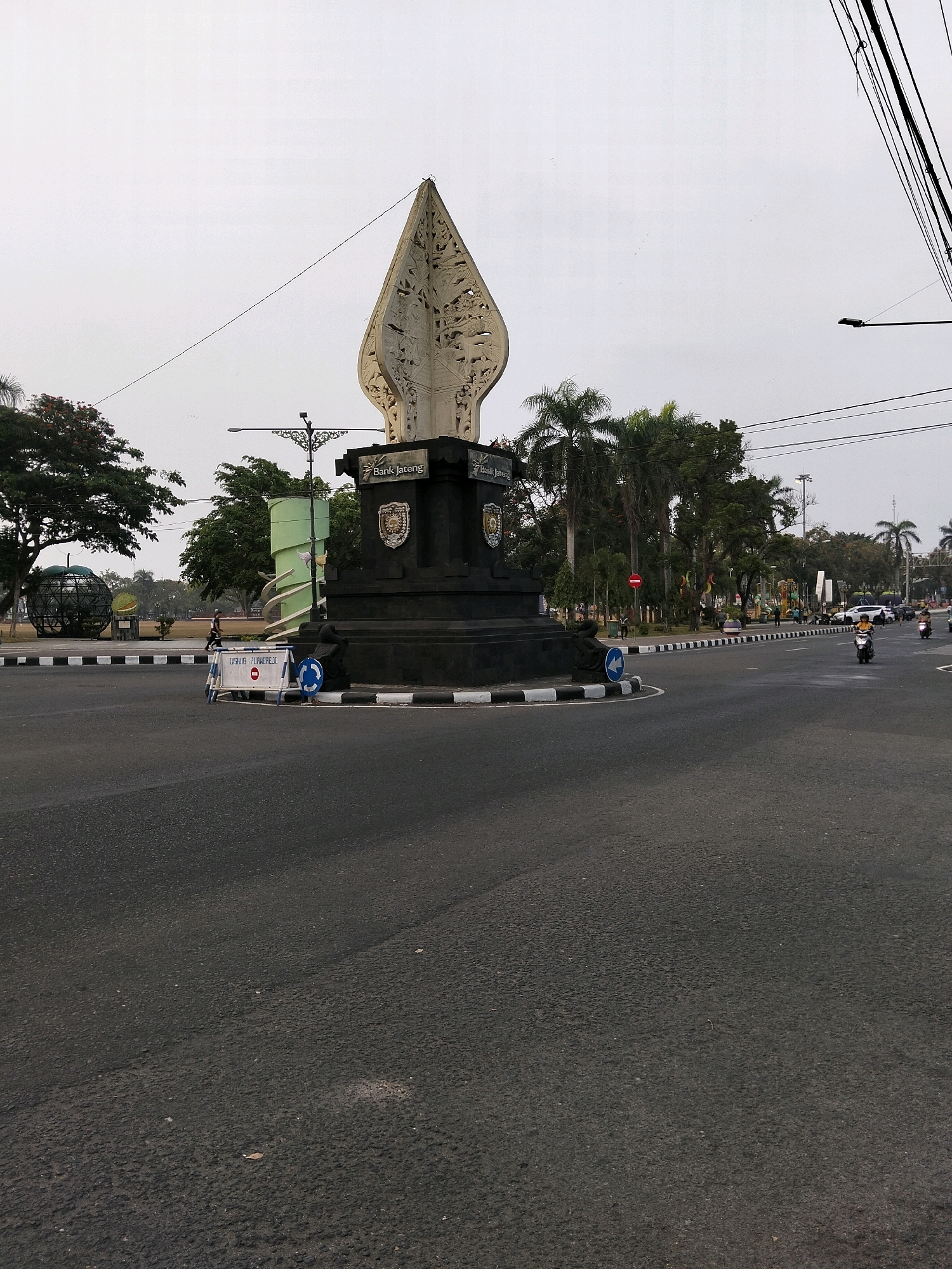The image captures a roundabout in Purworejo, Indonesia, dominated by a tall, abstract monument resembling a traditional Javanese 'keris' (dagger) or 'gunungan' (mountain shape from Wayang Kulit performances). The monument is dark and sits on a raised, circular base with decorative patterns. The sky is overcast, suggesting an early morning or late afternoon scene.
In the foreground, a wide asphalt road curves around the roundabout, marked with black and white stripes along its edge. Several small, round, blue and white traffic cones are strategically placed on the edge of the road, along with a white barrier with a red circular sign indicating a prohibition.
To the left of the roundabout, a large, spherical, metallic sculpture with an intricate geometric design is visible, along with lush green trees. Further back, a green, spiral-shaped structure stands out, and in the background, palm trees and other foliage indicate a tropical environment.
Several people and vehicles are present. A person is walking on the sidewalk to the left of the roundabout. On the road, several motorcycles are visible, with one rider wearing a yellow shirt and another in darker clothing. Cars are also present in the distance, moving along the road. The scene appears to be a public space within the city, likely a significant landmark given the prominent monument. The overall impression is one of a moderately busy urban intersection with a cultural monument at its center.