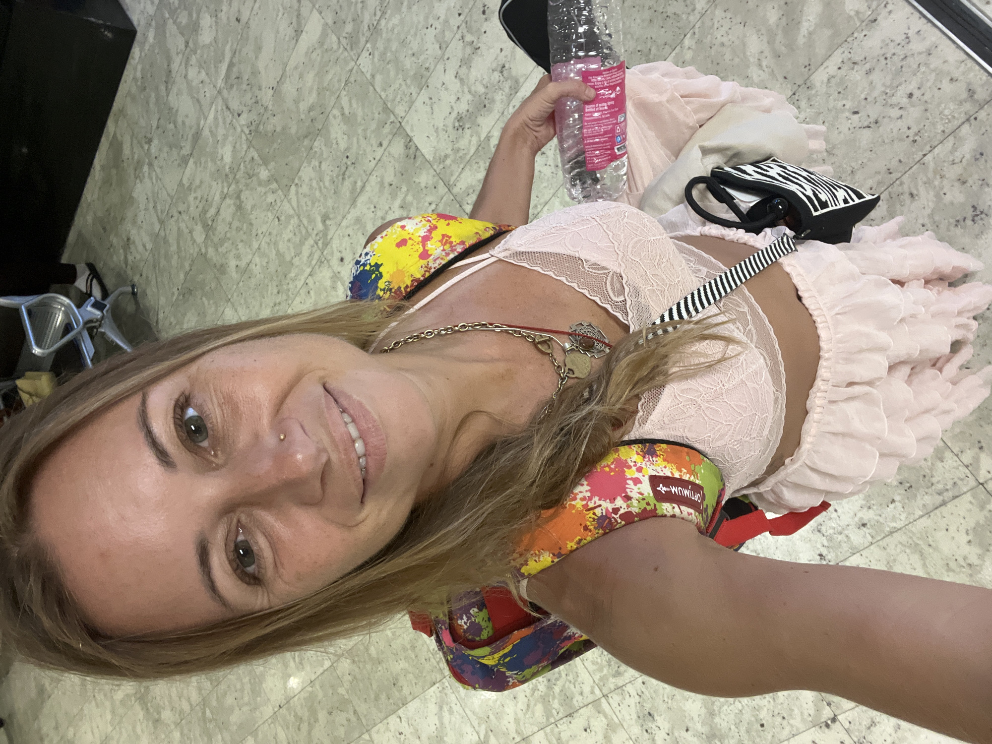 A woman is taking a selfie in Katunayake, Sri Lanka. She has blonde hair and is wearing a pink lace bra and a pink ruffled skirt. She is holding a bottle of water and has a colorful backpack on. The background appears to be an airport terminal with tiled flooring and seating.