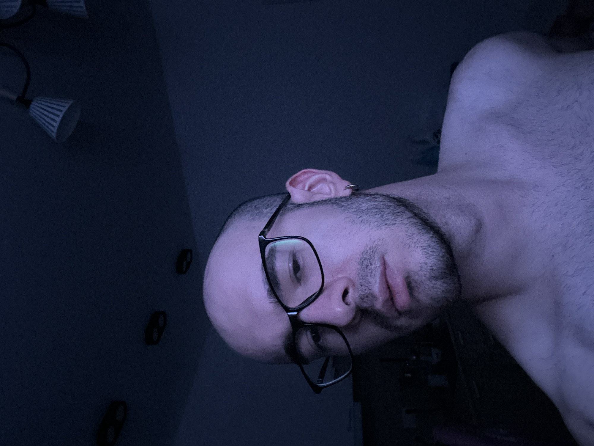 A low-angle close-up depicts a shirtless male individual with a bald head, dark beard, and glasses. The person's face is tilted slightly, with a neutral expression. The background is dark, showing a ceiling light fixture and three electrical outlets. The location is Unknown, Unknown.