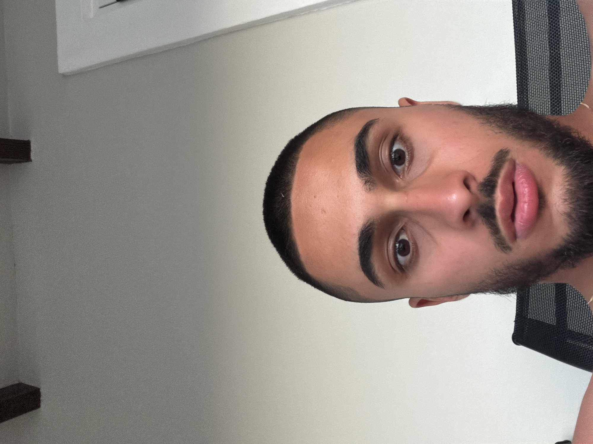 The image is a close-up, low-angle selfie of a young man with short, dark hair, dark eyebrows, and a short beard. He has brown eyes and is looking directly at the camera. His lips are slightly parted, and he appears to be wearing a necklace. He is seated in a black office chair with a mesh back. The background consists of a plain, light-colored wall and a dark wooden beam near the top of the frame. The lighting suggests an indoor setting, possibly a home or office. There is no discernible information within the image to indicate a specific location in Laganas Municipal Unit, Greece.