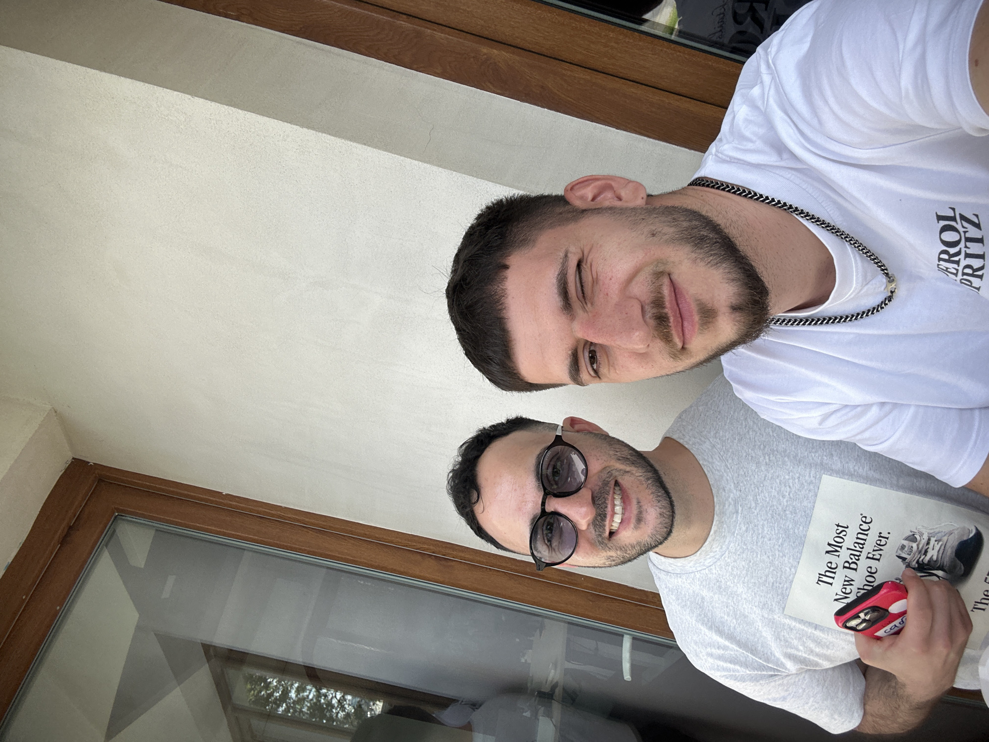 Two men pose indoors. The man on the top wears a white t-shirt with a chain necklace. The man on the bottom wears sunglasses and a gray t-shirt. He holds a book titled "The Most New Balance Shoe Ever" and a red object in his hand. A window frame is visible. The location is Sofia, Bulgaria.