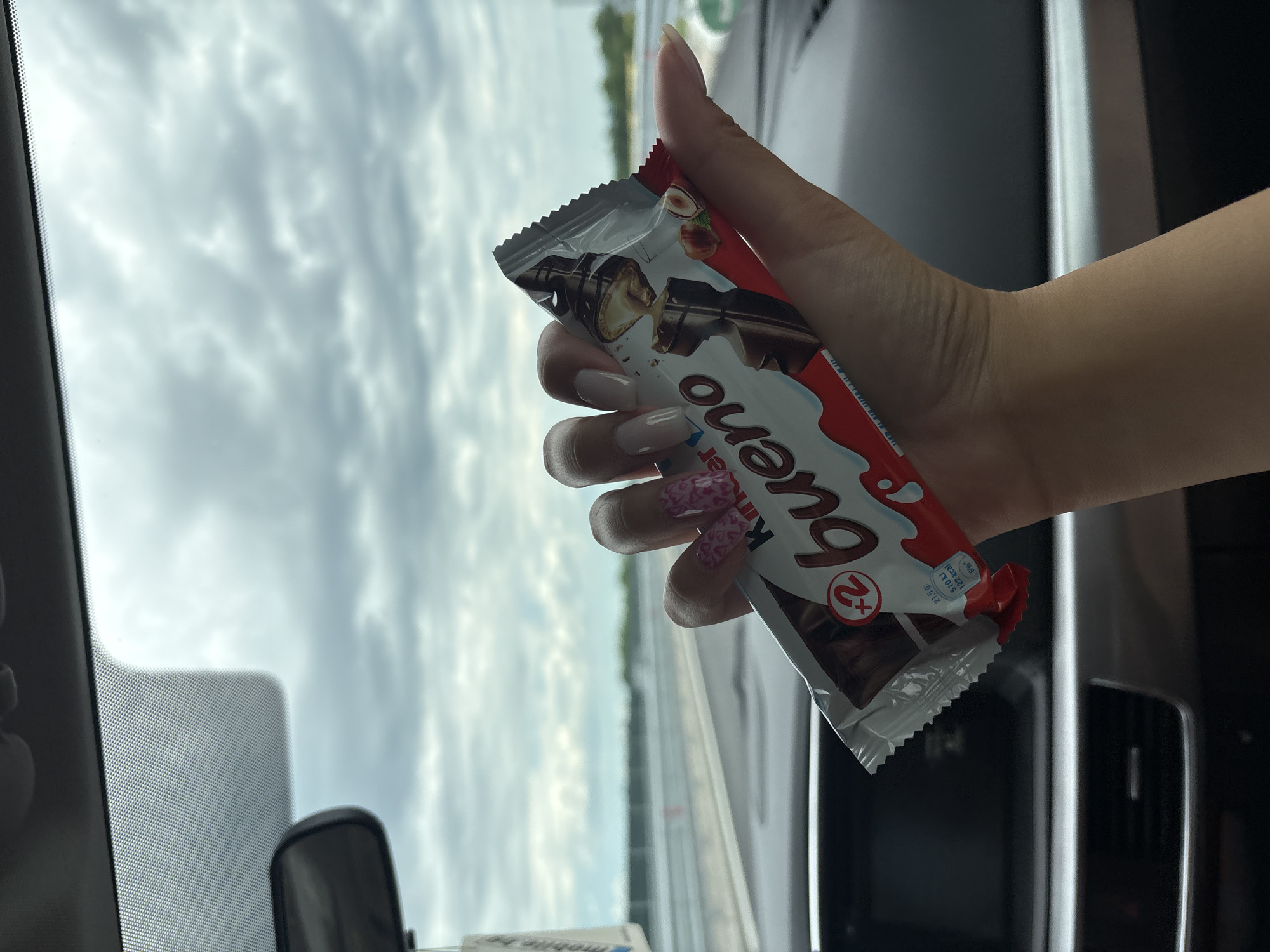 A person is holding a "Kinder Bueno" chocolate bar in their hand inside a car. The background shows a cloudy sky visible through the car window and a road or overpass. It is difficult to ascertain the location within the city of Peshterna, Bulgaria, without additional information.