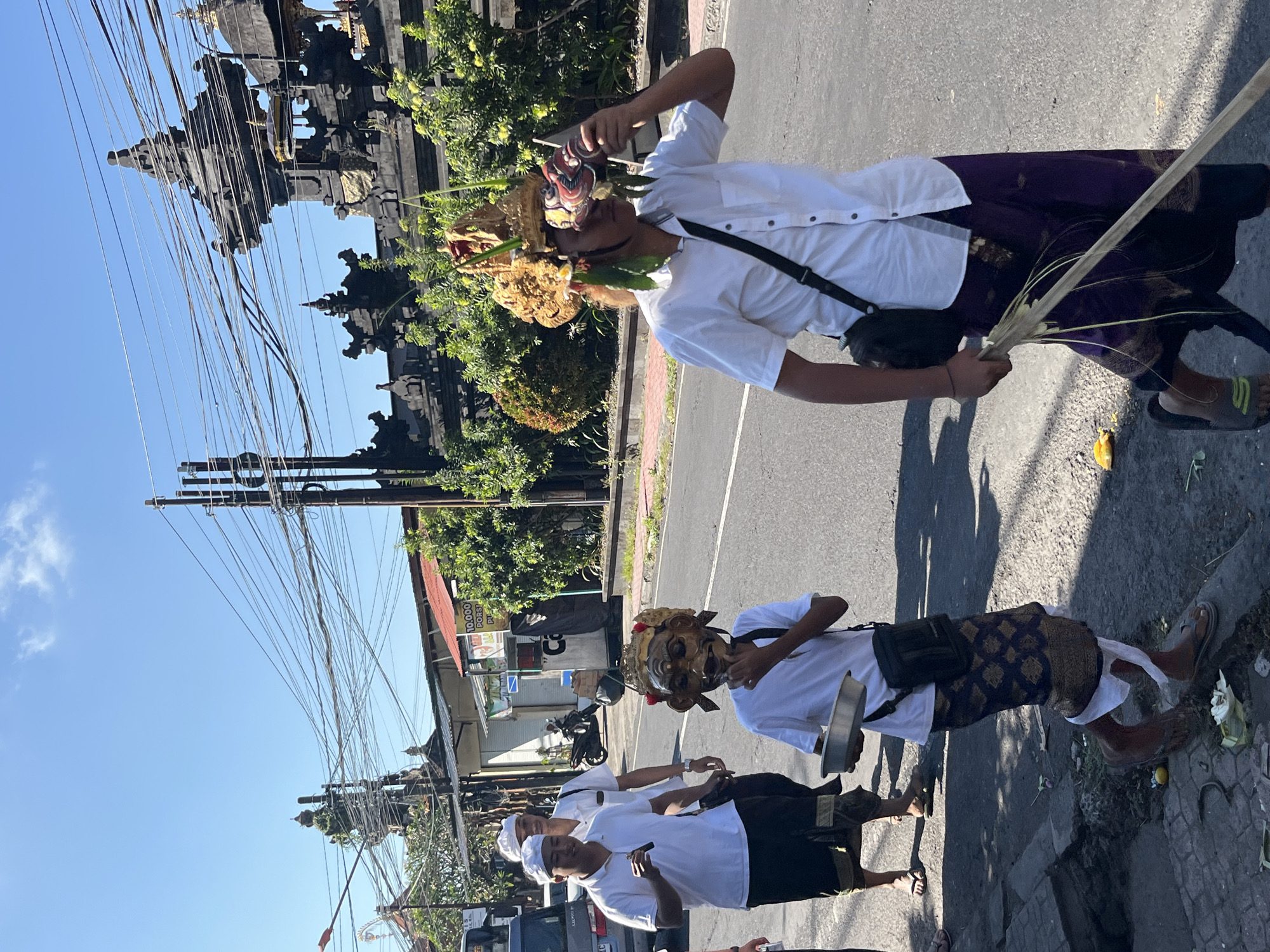 In Sukawati, Indonesia, under a bright blue sky, a procession moves along a paved street lined with greenery and traditional Balinese architecture. Two individuals are the primary focus. The person in the foreground, wearing a white short-sleeved shirt and a deep purple sarong with gold accents, carries a long, rustic wooden stick and a black crossbody bag. This person is adorned with an elaborate, golden headdress featuring masks and decorative elements, and holds up a small, colorful mask to their face, seemingly examining it.
Behind this individual walks another person dressed in a white polo shirt and a dark sarong, wearing a white cap. A third person, partially visible in the background, also wears a white shirt and a white cap. The street shows signs of activity, with a scooter parked in the middle ground and various buildings and utility poles with a dense network of wires visible. The environment suggests a warm, sunny day, possibly during a cultural event or ceremony.