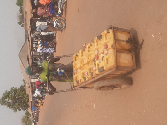 The scene depicts a street in Gwadanye, Nigeria. A dark-skinned adult male, wearing a light green sleeveless top and a dark blue/green headwrap, is pushing a two-wheeled metal utility cart down an unpaved, reddish-brown dirt road. The cart is loaded with at least twelve yellow plastic jerry cans, each fitted with a colored screw cap (some red, some white). In the background, numerous people are visible on both sides of the road, congregating near various single-story structures with corrugated metal roofs. Several motorcycles are present, some parked, others ridden by individuals. A prominent green tree is visible on the left side of the road in the distance. The environment suggests a busy, possibly market or commercial, street under clear daytime conditions.