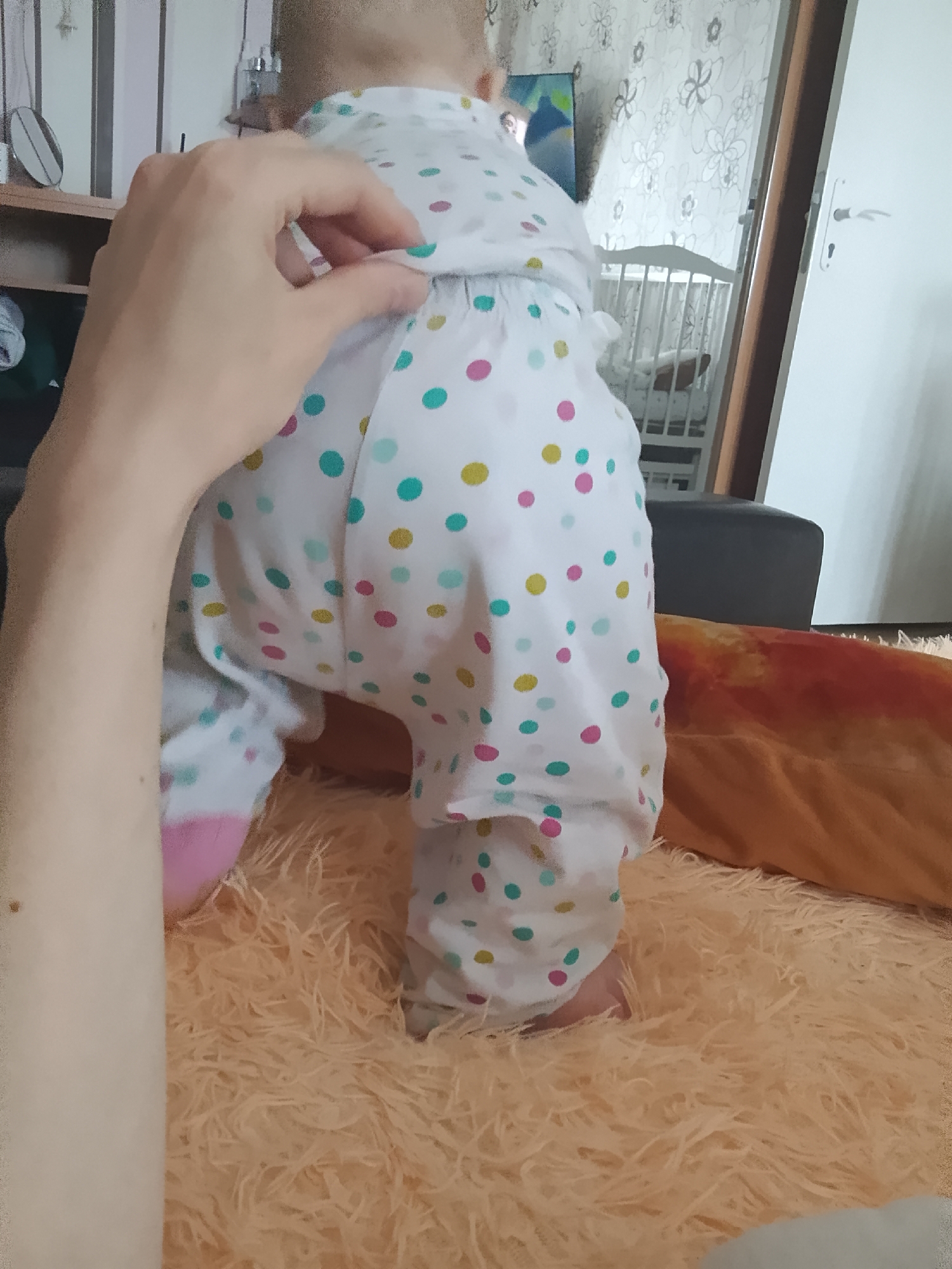 The image depicts a close-up view of a toddler standing on a fluffy, peach-colored rug. The toddler is wearing a white onesie adorned with colorful polka dots in pink, teal, and yellow. A person's hand is visible in the foreground, gently adjusting the toddler's clothing. In the background, a white crib with a railing is visible, suggesting a nursery or child's room. A portion of a white door with a silver handle is also present on the right side of the frame. The overall environment appears to be indoors, likely a residential setting. There is no identifiable information that places the scene specifically in the city of Minsk, Belarus.