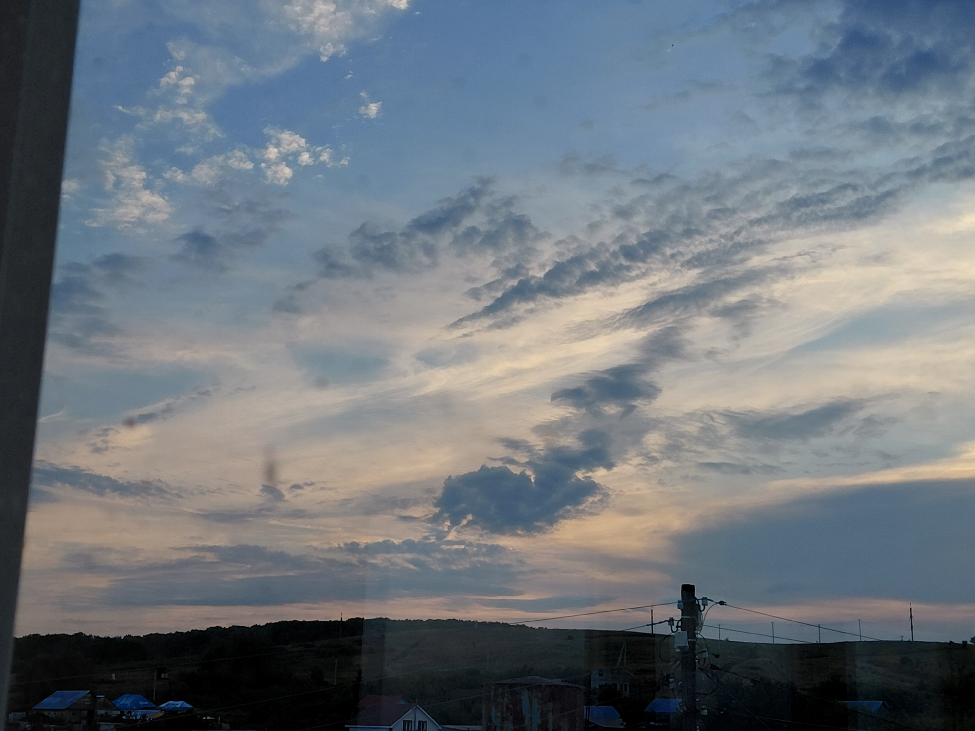 The image depicts a sky with scattered clouds, some dark and others light, against a pale blue and orange gradient, suggesting either dawn or dusk. Below the sky, a dark, undulating landscape of hills is visible, dotted with trees and some human settlements. Several houses with blue roofs and some industrial structures are discernible in the lower part of the image. Electrical poles and wires are also present, indicating the presence of infrastructure. The scene appears to be a rural or semi-rural area on the outskirts of Novokuybyshevsk, Russia, likely captured from a window or elevated vantage point. There are no people or explicit actions visible in the frame.