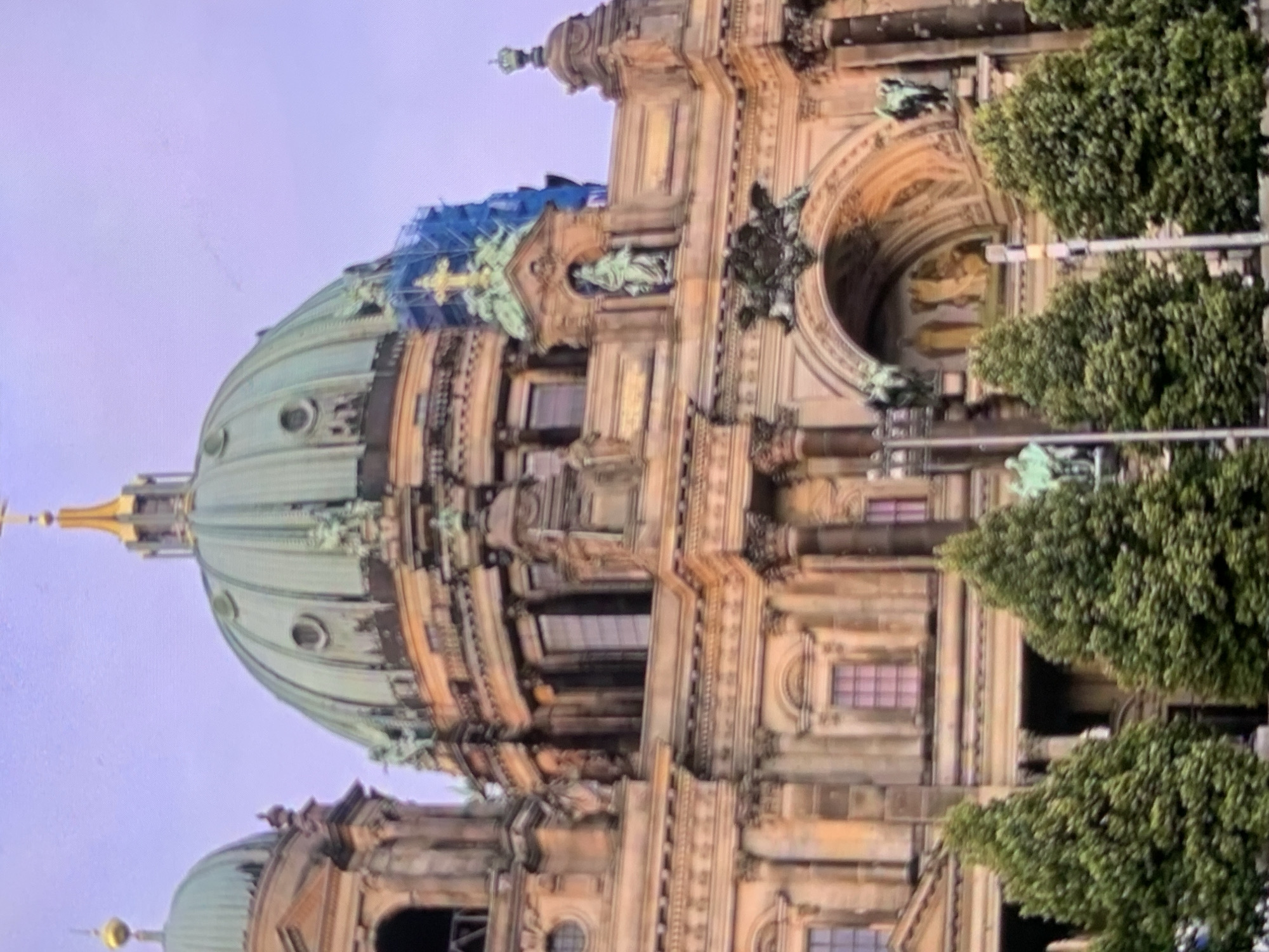 The image shows a close-up of the Berlin Cathedral, a large Protestant cathedral in Berlin, Germany. The cathedral features a prominent green dome, ornate architecture, and sculptures. Several evergreen trees are visible in the foreground, partially obscuring the lower part of the building. The sky above is a pale purple, suggesting it might be dusk or dawn. There is some scaffolding and blue netting visible on one of the upper sections of the building, indicating ongoing construction or maintenance. The scene is a static, architectural shot without any people or overt action. The location is identified as Stavropol, Russia, which is incorrect as the building is in Berlin, Germany.