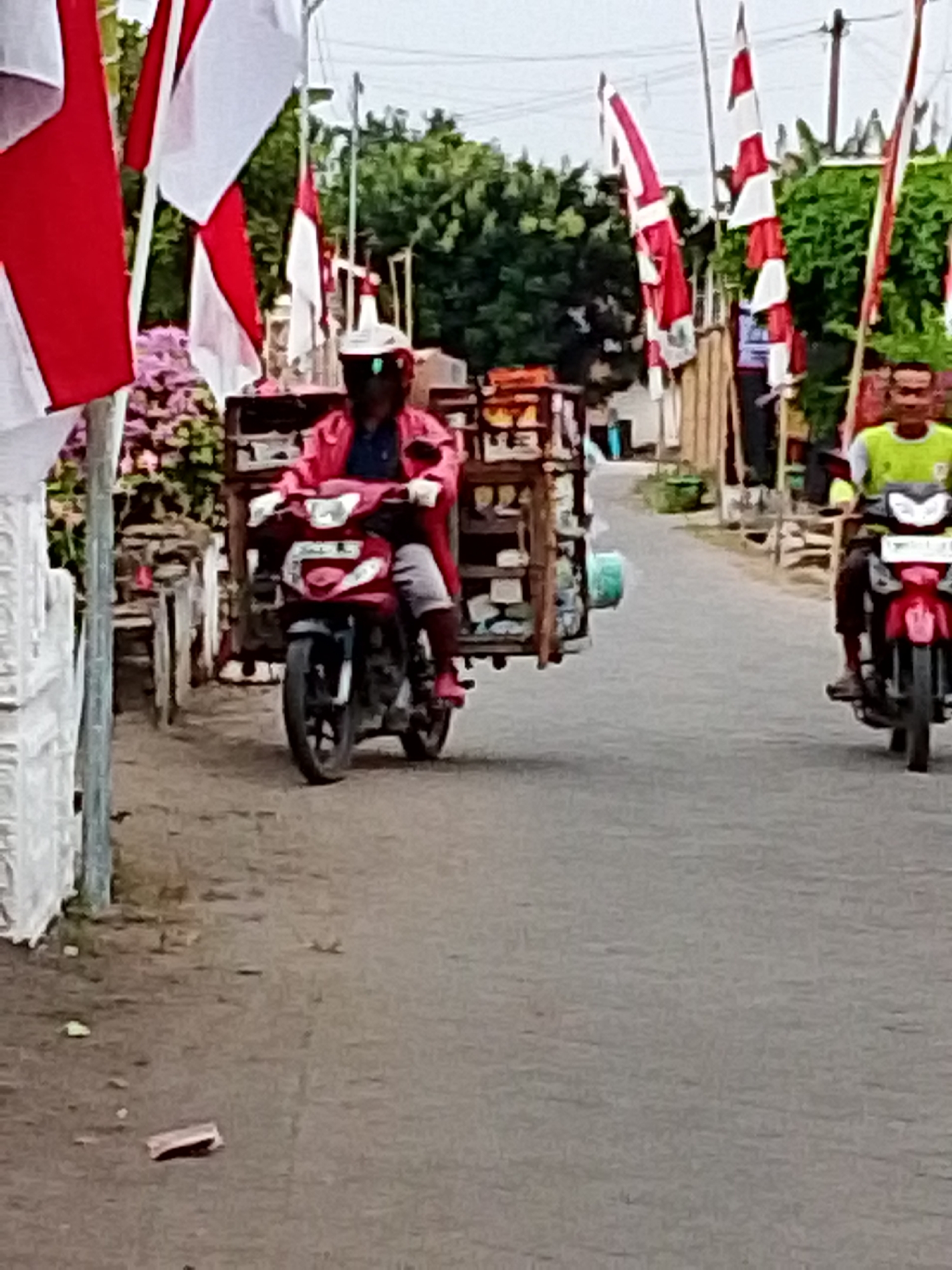 In Bulurejo, Indonesia, two individuals are depicted riding motorcycles on a paved road. The foreground shows a person on a red scooter, wearing a pink jacket and a white helmet. Behind them, attached to the scooter, is a wooden cart laden with various items. To the right, another individual is riding a red motorcycle, wearing a bright green shirt and a helmet. The background features lush green trees, and several Indonesian flags, characterized by their red and white stripes, are prominently displayed on poles along the roadside. The atmosphere suggests a local scene, possibly during a celebration or event, given the flags.