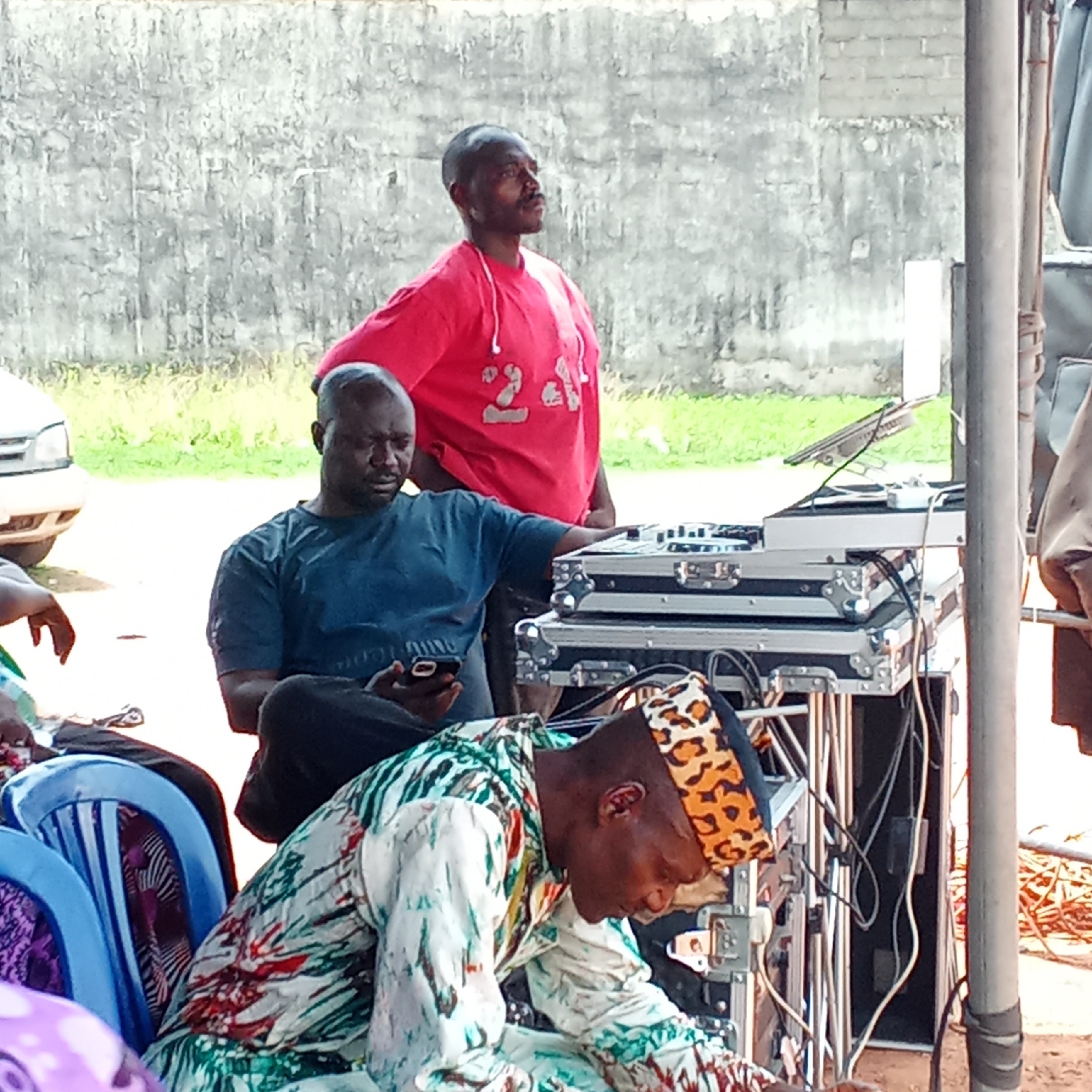 This image depicts an outdoor scene, likely an event or gathering, taking place in Akwanga, Nigeria, during the daytime. Three men are prominently featured.
In the lower right foreground, a man with a dark complexion is shown bending or crouching. He wears a vibrant, multicolored patterned long-sleeved outfit and a distinctive leopard-print cap, and appears to be attending to the cabling of DJ equipment. Immediately behind him, another man with a dark complexion sits on a chair, engrossed in his mobile phone. He is dressed in a dark blue t-shirt and dark pants, with his arm resting on the adjacent DJ setup. Standing to their right and slightly behind is a third man, also with a dark complexion, wearing a red long-sleeved t-shirt with a faint white design (possibly "23"). He has short hair and a mustache, and is looking off to the right with an observant expression.
The central focus of the scene, besides the individuals, is the DJ equipment. It consists of two silver, flight-case-style boxes stacked on a stand, topped with a laptop placed on a separate raised stand. A network of wires and cables connects the various components. The background features a light gray, textured concrete block wall and a strip of green vegetation. A portion of a white car is visible on the far left, along with blue plastic chairs. Vertical metal poles on the right suggest the presence of a temporary structure or canopy under which the event is being held. The ground beneath the participants appears to be dirt or sand. No clear, legible text is visible anywhere in the image.