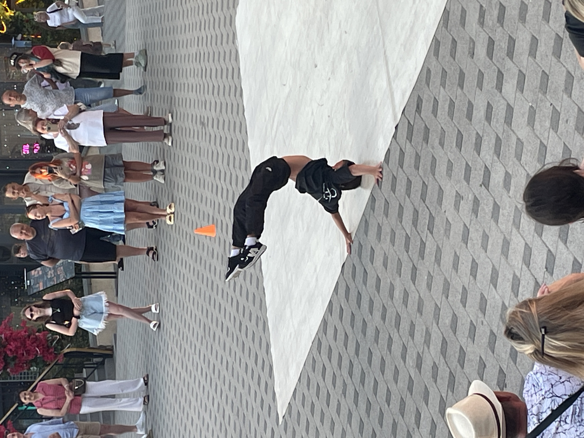 In an outdoor, urban setting with patterned grey paving stones, a young person is performing a breakdancing move, balancing on their hands with their legs extended upwards. They are wearing a black t-shirt and black pants. A white mat is laid out on the paving stones for the performance. In the background, a crowd of people, dressed in casual attire, watches the performance. Among the onlookers are several individuals of various ages and genders, some holding phones, possibly to record the event. A bright orange cone is visible on the paving stones to the right of the breakdancer. The environment appears to be a public space, possibly a plaza or walkway, with some foliage and architectural elements visible in the distance.