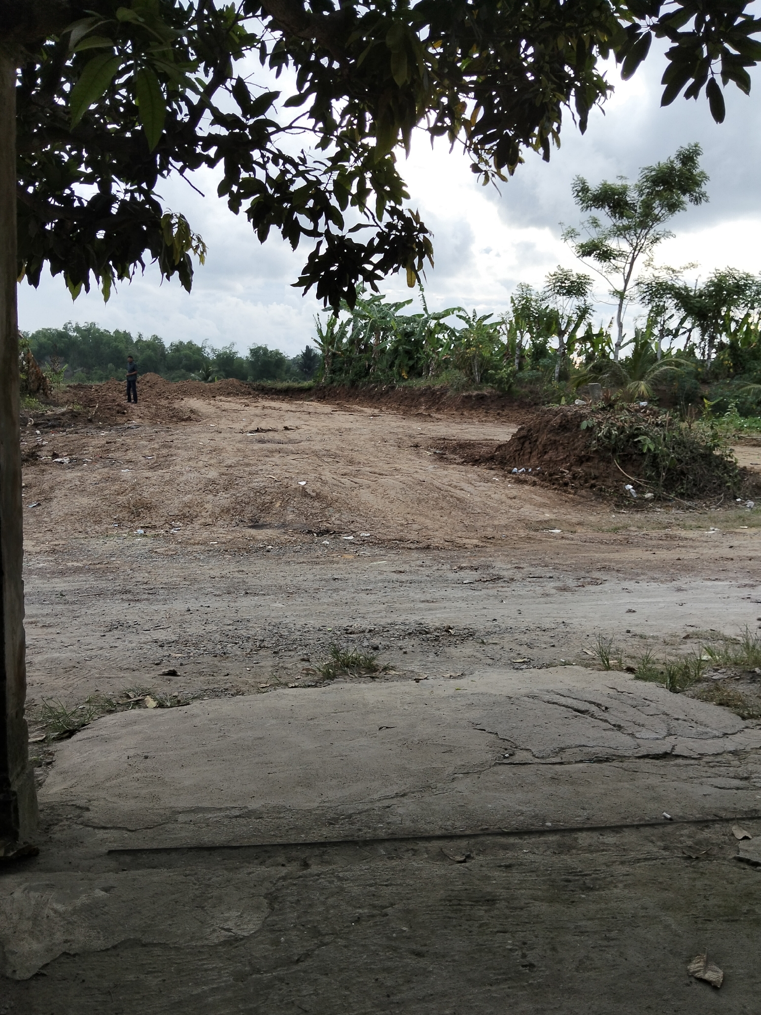 The image shows a construction site in Payagaboh, Indonesia, with a pile of dirt in the middle ground. A single person, appearing to be a male in a dark shirt and pants, stands on the dirt pile in the left-center of the frame. The background is filled with lush green vegetation, likely banana trees, and a dense line of trees in the far distance. The foreground is a rough, concrete surface with cracks and small stones. The sky is cloudy and overcast. The scene suggests ongoing outdoor work or land preparation.