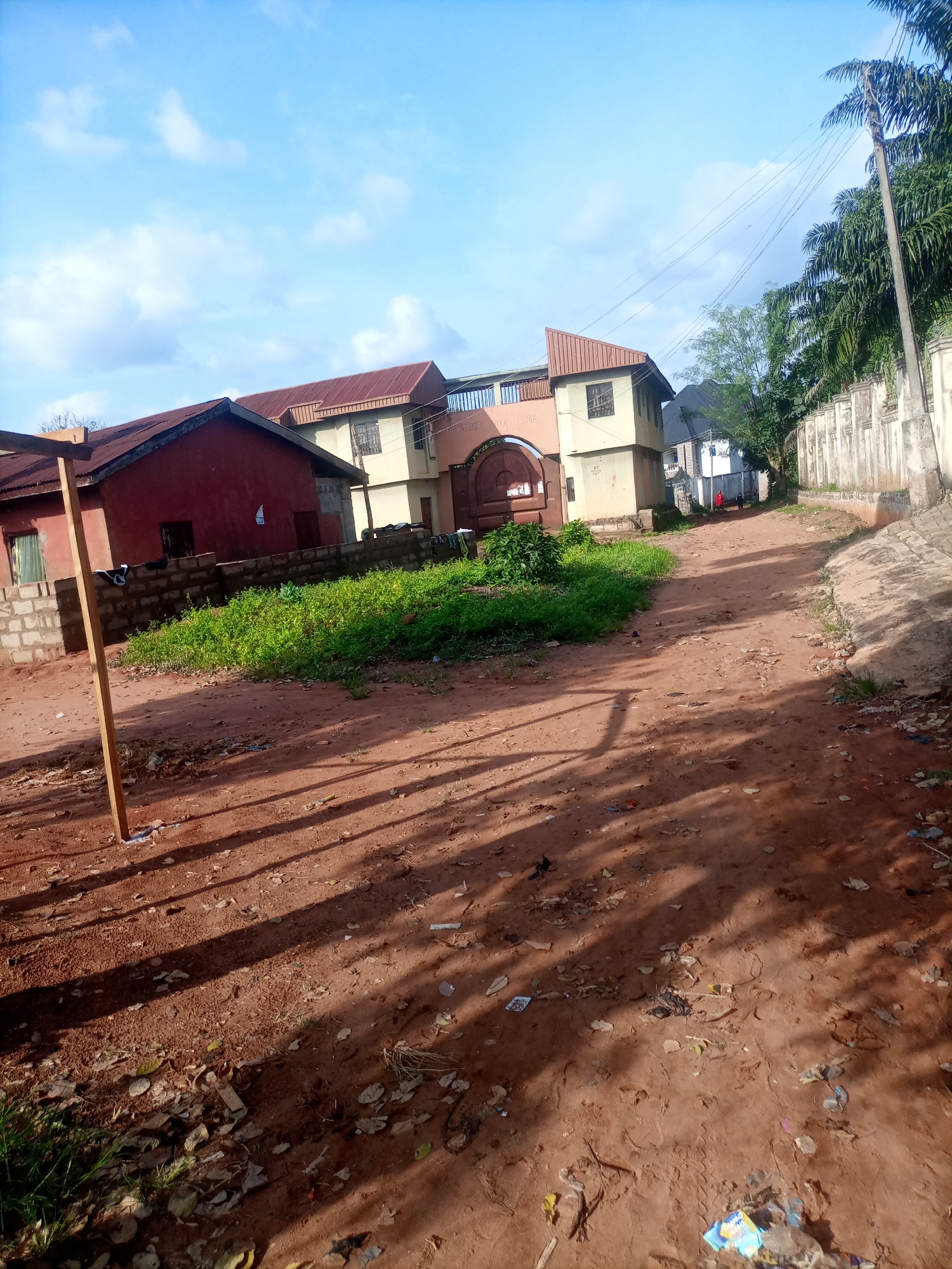 The image depicts a dusty, red-earth road in Nkpor, Nigeria, leading towards a cluster of buildings. The road is sparsely littered with debris. On the left side of the road, a low brick wall encloses an area with vibrant green grass. Behind this wall, a red-colored building with a corrugated metal roof stands prominently. To the right of this red building, a beige multi-story structure with a similar metal roof is visible. Further down the road, more buildings and a tall concrete wall with decorative elements can be seen. Palm trees and other foliage line the right side of the road, suggesting a tropical or subtropical environment. The sky above is a clear blue with scattered white clouds. The lighting suggests it is either early morning or late afternoon, casting long shadows across the road. No people are clearly visible in the immediate foreground, though distant figures might be present further down the road. The overall scene conveys a sense of a residential or community area in an African city.