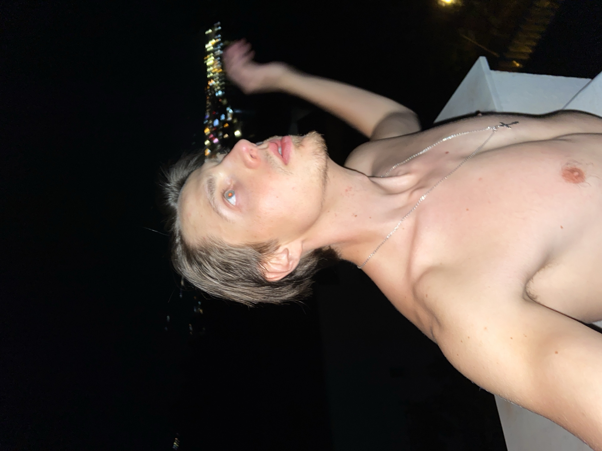 A shirtless male stands at night on a white concrete ledge, with a cityscape illuminated in the background. The man has blond hair, a light beard, and a silver chain with a cross pendant on his chest. His right arm is extended upward, partially obscuring the cityscape. The location is undetermined.