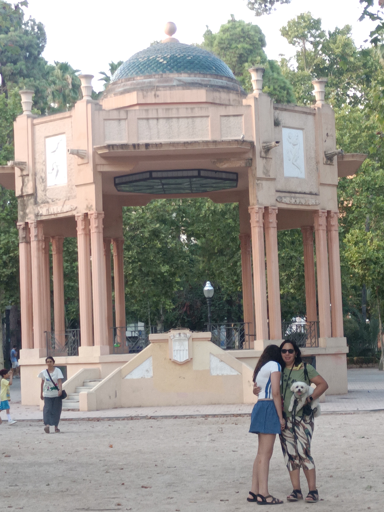 The image depicts a sunny day at the Parque del Oeste in Castelló de la Plana, Spain. In the foreground, two women are embracing. The woman on the left, wearing a white shirt with blue stripes and a blue skirt, has her back to the camera. The woman on the right, wearing a green t-shirt and camouflage-patterned capri pants, is holding a small white dog in her arms. She has dark hair and is wearing sunglasses. Behind them, a woman in a white shirt and dark skirt walks away from the camera, carrying a black bag over her shoulder. A young boy in a yellow t-shirt and blue shorts walks in the opposite direction. In the background, a domed pavilion with decorative pillars stands amidst lush green trees. The ground is a light-colored sand or gravel.