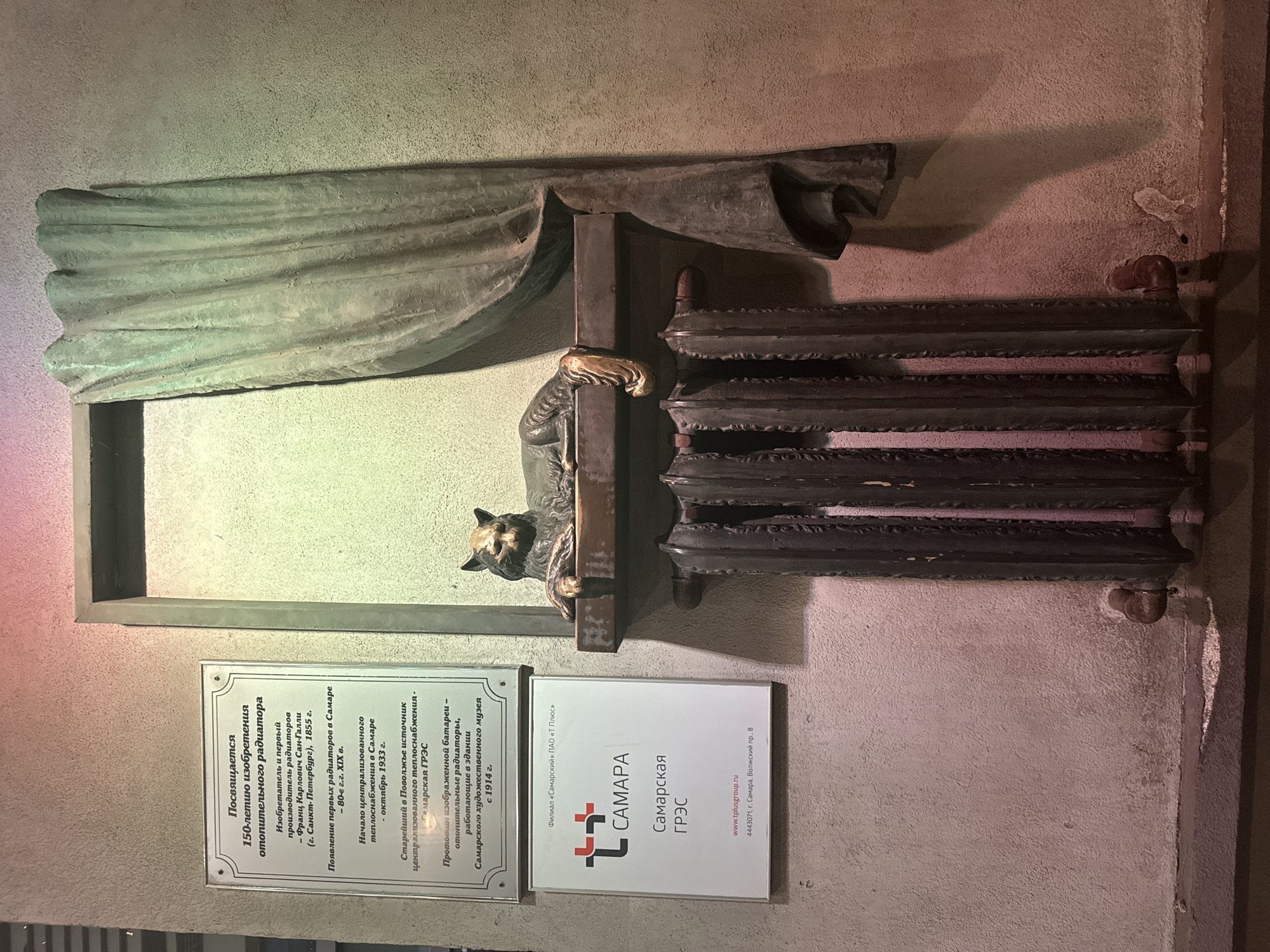 The image depicts an outdoor sculpture installation in Samara, Russia, dedicated to the invention of the heating radiator. The sculpture features a bronze cat resting on a radiator, with a draped curtain on the right side. A plaque above the sculpture details the history of the radiator, noting its inventor and the introduction of centralized heating in Samara. Below the plaque, a sign for "Samarskaya GRЭС" (Samara State Regional Electric Station) is visible, along with their website and address. The sculpture is mounted on a textured concrete wall, illuminated by ambient lighting that casts shadows. The installation appears to be a public art piece, likely a monument or a decorative element in a public space.