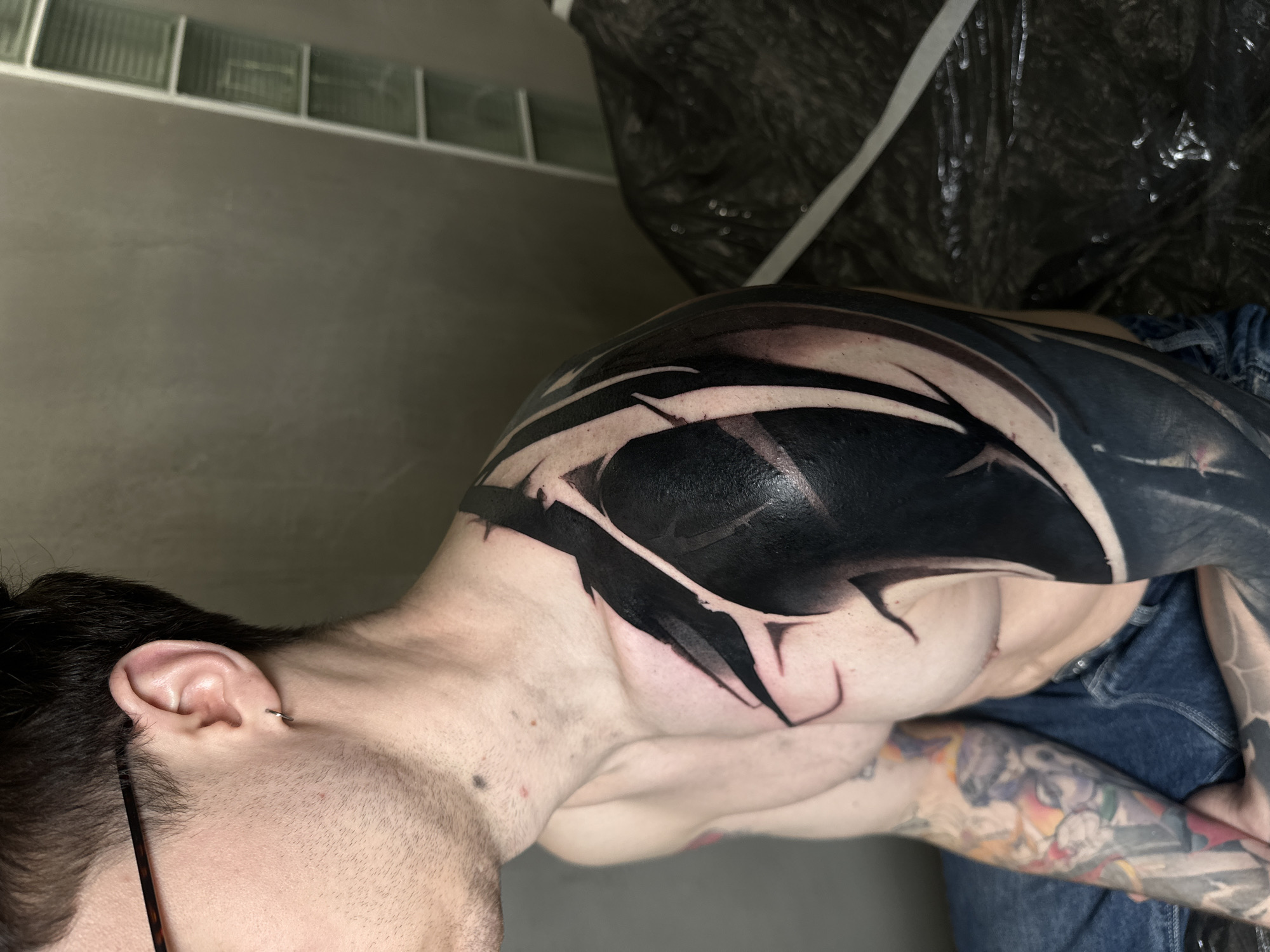 A close-up, high-angle shot depicts a shirtless man from the neck down, displaying extensive tattoos. The primary tattoo features a large, abstract black design on his left shoulder and upper chest, accented with white lines. A smaller, colorful tattoo is visible on his left forearm. He is wearing blue jeans. The background consists of a grey wall with glass bricks near the top, and a dark, draped plastic-like material. The location and city information are not evident from this image.
