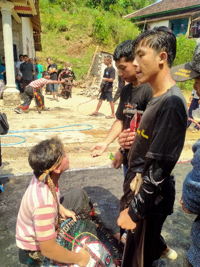 The image depicts an outdoor cultural performance in Ngargoyoso, Indonesia, under bright sunlight. In the immediate foreground, a young male individual kneels on a damp, dark ground surface. He wears a striped pink and white short-sleeved shirt, dark pants, and a patterned headscarf, with disheveled hair showing traces of yellow substance. He holds a large, colorful, circular object resembling a woven horse head prop, part of a traditional Kuda Lumping costume, and gazes upwards with an open mouth. Directly in front of him stand two young men in wet black short-sleeved shirts. The man on the right holds a string of dark beads (tasbih) and extends an open left hand towards the kneeling individual's mouth. His companion stands beside him.
In the mid-ground, to the left, a performer in a dark, horse-like costume (Kuda Lumping) extends their arm towards another individual holding a whip. A blue hose lies coiled on the ground. To the far left, a white-pillared building with a tiled roof is partially visible, with a group of onlookers gathered nearby. The background features a steep, verdant hillside covered in dense vegetation, and a simple house with a light blue facade on the right. The ground is primarily dry earth, with localized wet areas.