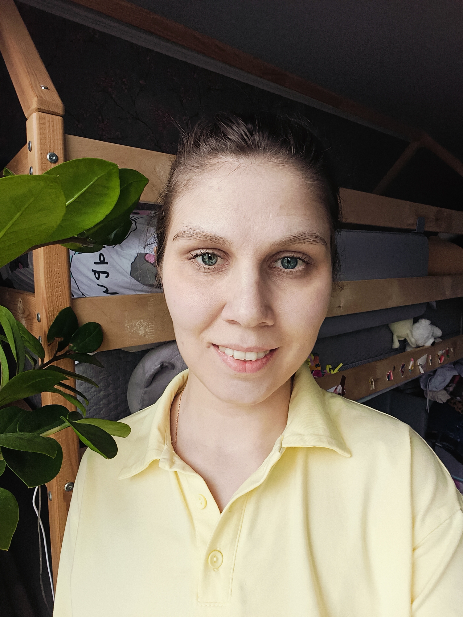 A young woman with light skin, blue eyes, and dark hair pulled back is smiling at the camera. She is wearing a yellow collared shirt. To her left is a green plant with large, glossy leaves. Behind her is a wooden bunk bed with gray bedding and various objects on the bottom bunk. The location appears to be indoors, possibly a bedroom. The city and exact location are unknown.