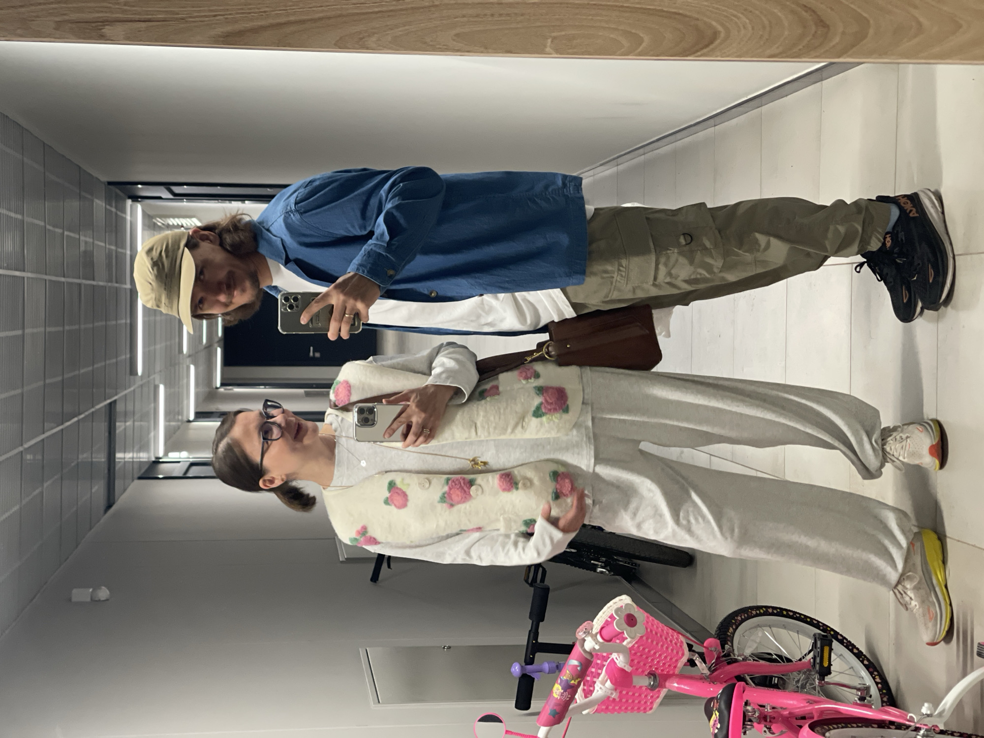 A mirror selfie captures two individuals in an indoor hallway. In the background, a person with long, dark hair under a light brown baseball cap wears a blue long-sleeved shirt over a white t-shirt, olive green cargo pants, and black athletic shoes with "HOKA" visible on the sole and "SKY" on the sock. This person holds a silver-cased smartphone in their right hand, taking the picture, and carries a brown leather crossbody bag. They exhibit a subtle smile.
In the foreground, a second person with dark hair tied back wears glasses, a white long-sleeved shirt, light grey loose-fitting pants, and white athletic shoes with yellow soles. They are adorned with a cream-colored vest featuring pink rose and green leaf appliqués, a thin necklace with a small pendant, and hold a silver-cased smartphone in their left hand. This individual also shows a gentle smile.
The setting appears to be a brightly lit, modern indoor corridor with white tiled flooring and linear light fixtures along the walls. A wooden trim is visible along the top edge of the mirrored surface. A pink child's bicycle with a front basket and streamers is partially visible at the bottom right of the frame, reflected behind the second person. Both subjects appear relaxed and content.