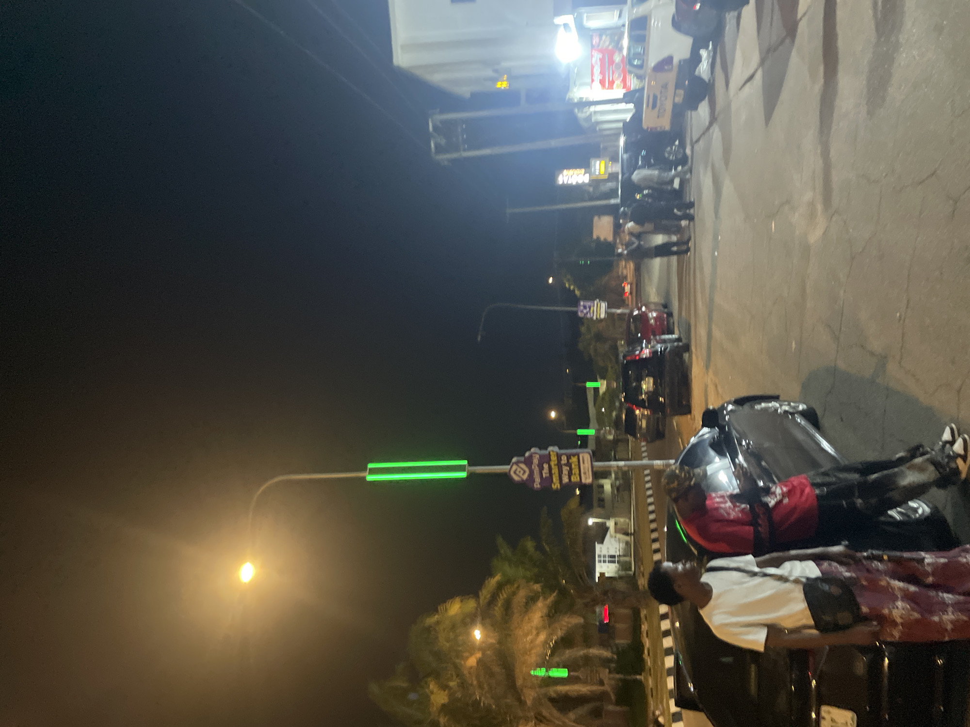 At night in Abuja, Nigeria, two individuals stand near a black car parked on a paved street. One person wears a white shirt, a black bag, and a patterned skirt, while the other sports a red shirt and dark pants. Streetlights illuminate the scene, including a lamp post with green lights along its length. Other cars and people are visible in the distance. Palm trees are also present.