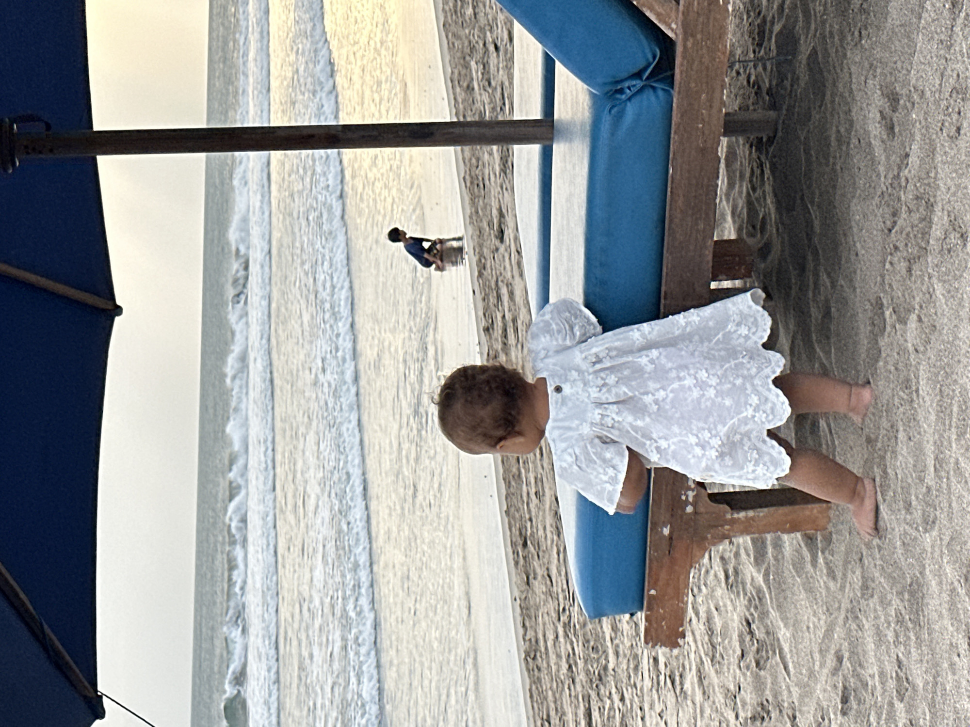 A toddler in a white lace dress stands on a sandy beach in North Kuta, Indonesia. The child is facing away from the camera and appears to be looking towards the ocean, where gentle waves are breaking on the shore. The sun is low in the sky, casting a warm, golden light on the scene, suggesting it is either early morning or late afternoon. The toddler is positioned next to a wooden beach chair with a blue cushion. In the background, another small figure, possibly a child, can be seen wading in the shallow water. The overall atmosphere is peaceful and serene, evoking a sense of calm and family enjoyment by the sea. There is no visible text in the image.