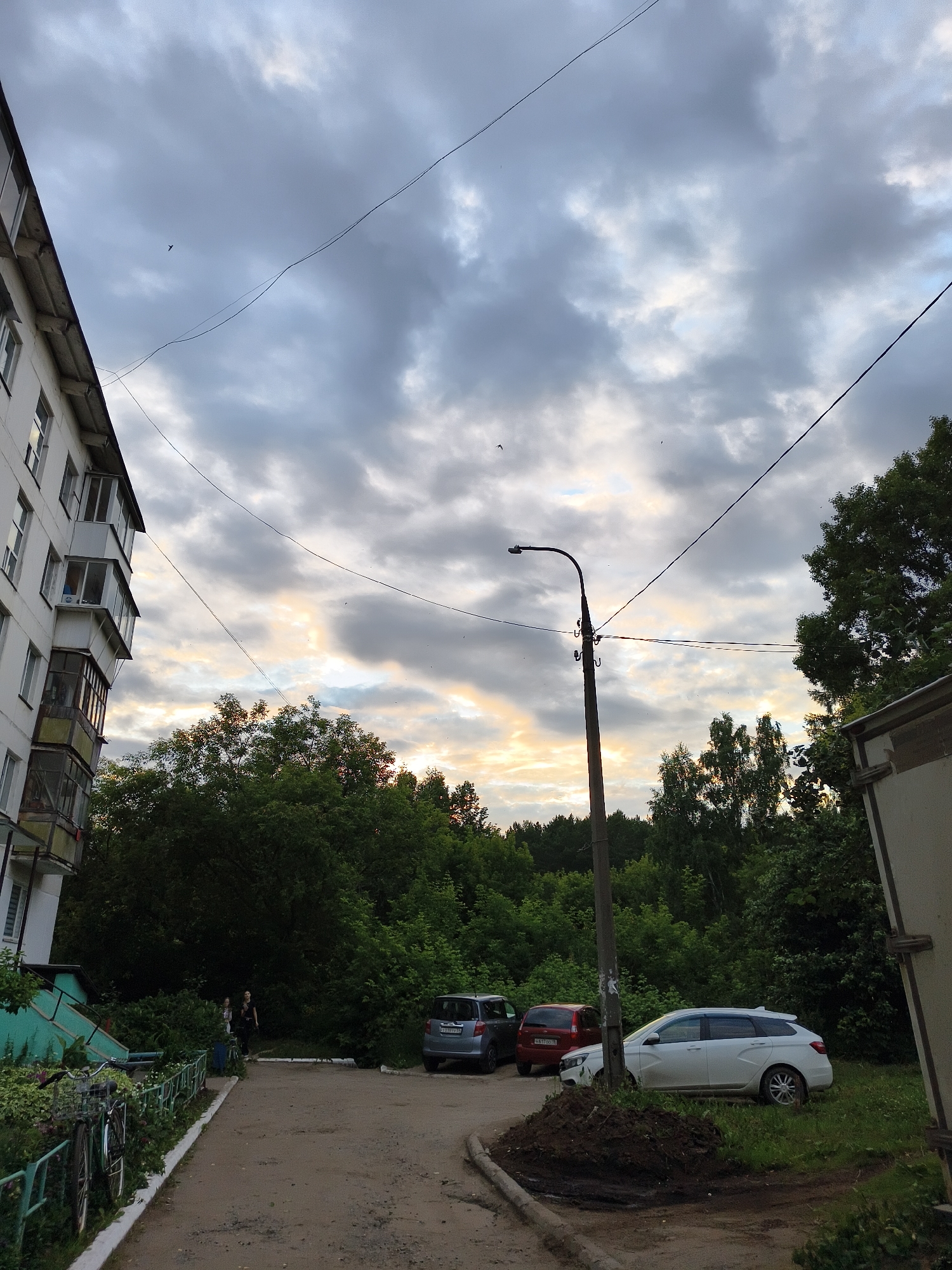 The image depicts a residential courtyard in the city of Chaikovsky, Russia, under a cloudy sky with streaks of yellow and orange light suggesting sunset. On the left, a multi-story apartment building with white facade and enclosed balconies dominates the scene. A paved walkway leads away from the building, bordered by a green metal fence with overgrown plants and two parked bicycles. Further down the walkway, two individuals, a woman in dark clothing and another person in lighter clothing, are walking.
The courtyard opens into a dirt and gravel area with parked vehicles. A silver sedan is parked on the right, partially on the grass. In the background, a dense line of green trees and foliage forms a backdrop, with a lamppost standing in the middle ground. Another car, red, is visible behind the silver sedan. The overall impression is of a typical urban residential area during late evening.