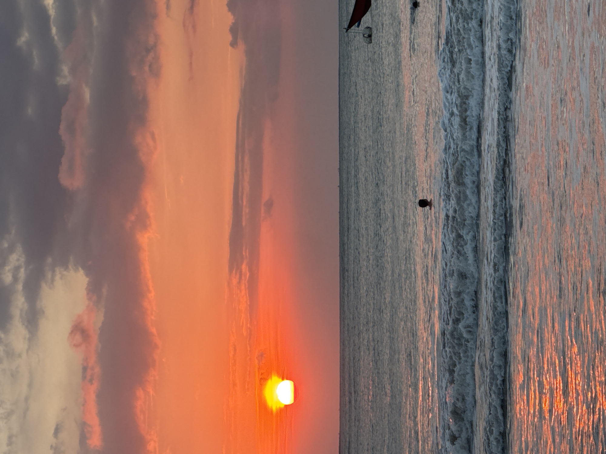 The image features a serene sunset over the ocean, likely in Kuta Selatan, Indonesia. The horizon line horizontally divides the frame, separating the vibrant sky from the reflective water.
The left side of the image is dominated by the setting sun, a brilliant yellow-orange orb low on the horizon, casting a warm, intense glow across the sky. Above and around the sun, the sky displays a gradient of fiery orange and soft pink hues, blending into darker grey and purple clouds at the top left. The clouds are scattered and textured, illuminated by the last rays of sunlight.
The right side of the image shows the ocean, with gentle waves breaking and receding towards the shore. The water's surface beautifully reflects the sky's colors, appearing as shimmering vertical streaks of orange and pink, particularly on the wave crests and wet sand in the foreground. A lone, silhouetted figure of a person is visible in the water in the mid-ground, appearing to be wading or swimming. Further out, near the horizon line, a small boat or watercraft with a dark, triangular sail or flag is faintly discernible.
The scene captures the peaceful beauty of an evening at the beach, characterized by the dramatic colors of the sunset and the tranquility of the ocean. There is no visible text in the image.