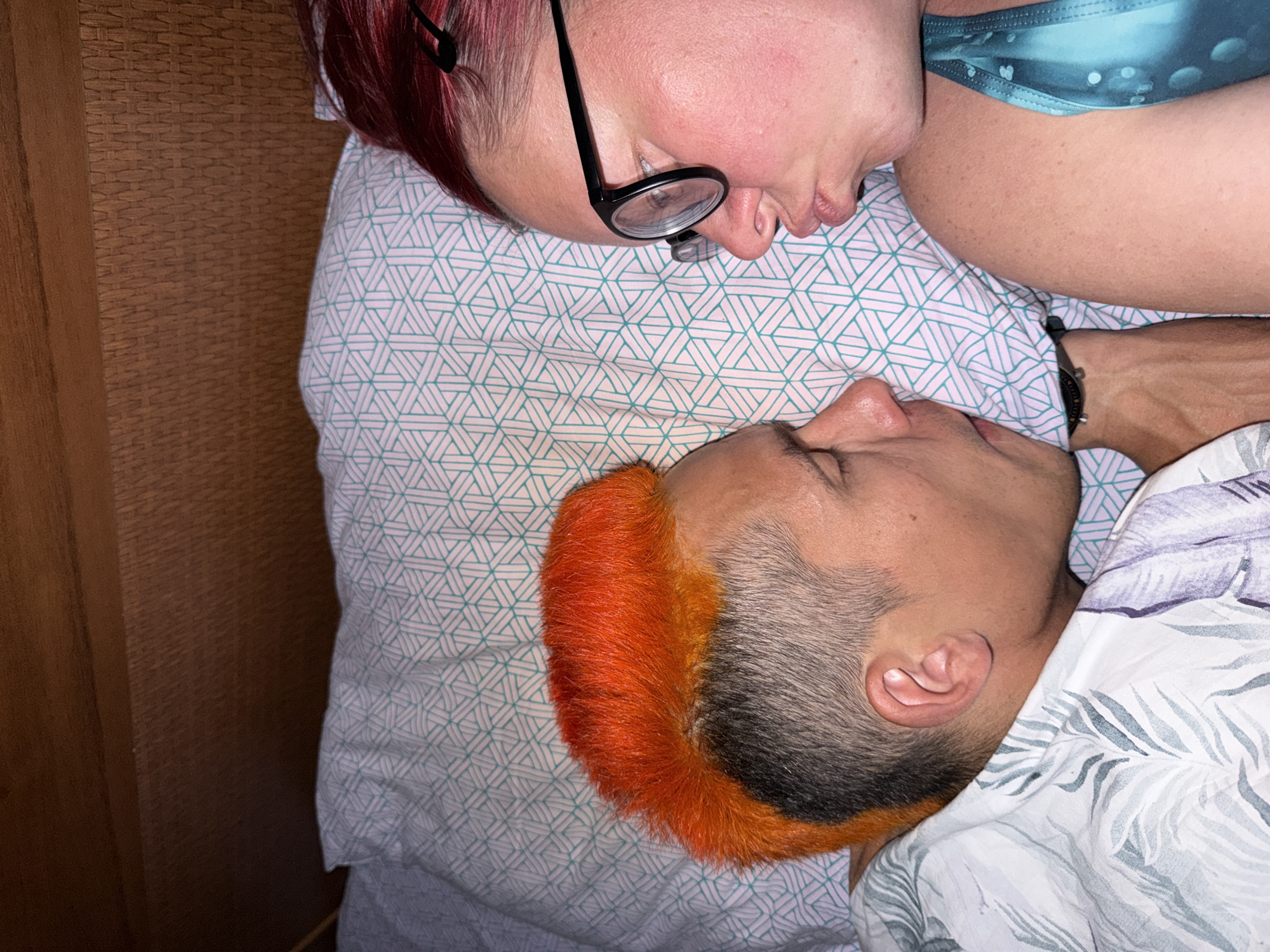 Two people are lying on a bed. One person has dyed red hair and is wearing glasses and a blue patterned top. The other person has orange and gray hair and is wearing a white patterned shirt. They are lying head-to-head on a light blue and white patterned pillow. The setting appears to be indoors, possibly a room in Chaikovsky, Russia.