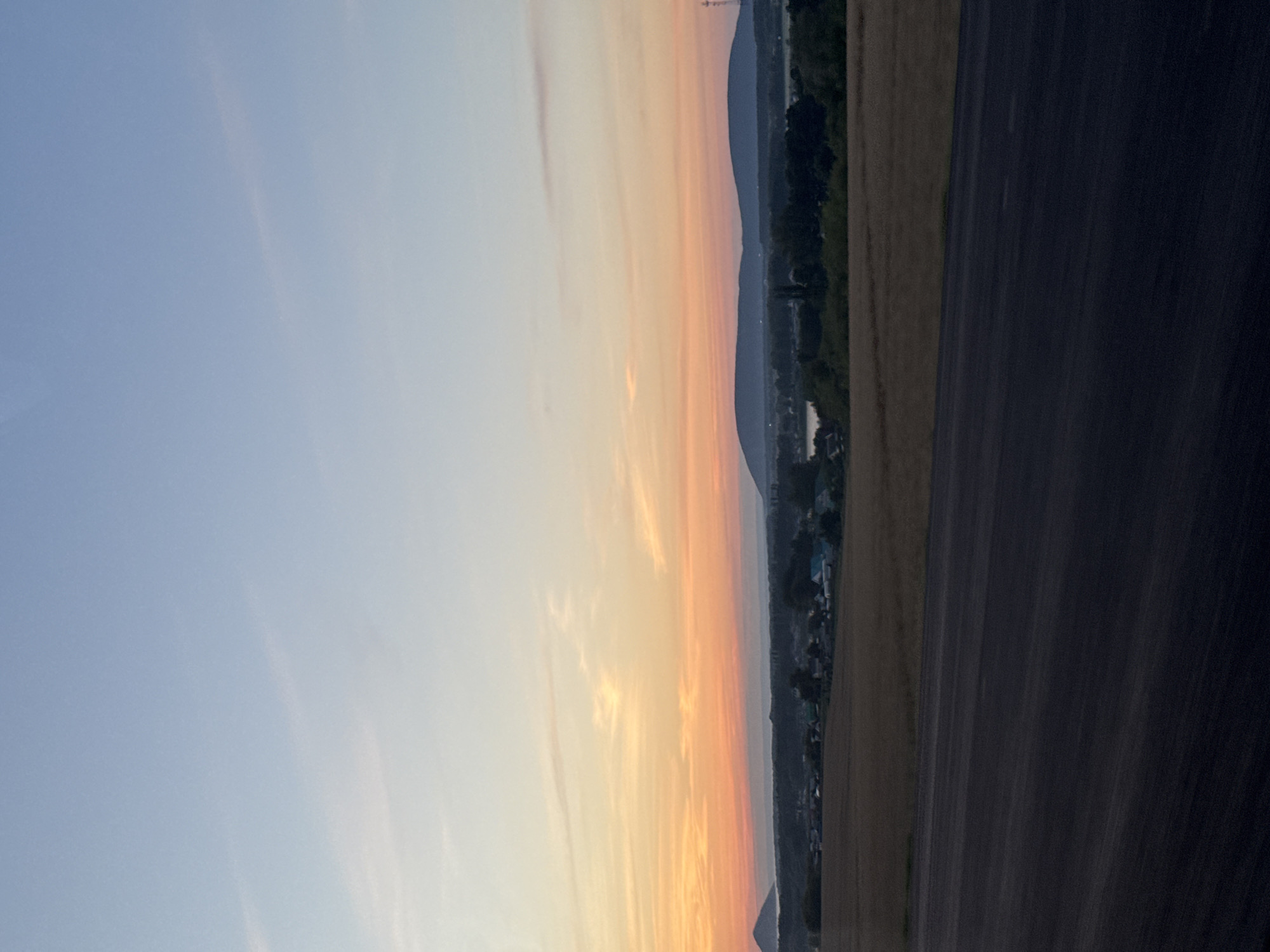 The image depicts a wide-angle, eye-level view of a rural landscape at dusk. In the foreground, a dark, textured asphalt surface occupies the lower portion of the frame. Beyond this, a field of dry, golden grass stretches horizontally. In the midground, a cluster of buildings, indicative of a village, is nestled amidst trees and a body of water. The village appears to consist of small houses with a mix of light-colored and blue roofs, and some trees with dark foliage. Further in the background, rolling hills with dark blue silhouettes form the horizon. The sky above is a gradient of colors, transitioning from a pale blue at the top to a soft orange and pink near the horizon, suggesting the setting or rising sun. There are faint, wispy clouds present in the sky. The overall scene conveys a sense of tranquility and natural beauty. The location is identified as the city of Muravei, Russia.