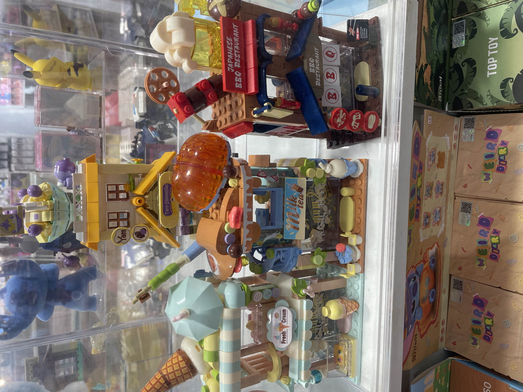 The image displays a collection of colorful, block-style toy models, resembling miniature buildings and food stalls, arranged on clear shelves within a display case. Prominently featured are ice cream cones, umbrella-shaped canopies, and signage indicating "Fresh Cream," "Taro Smoothies," "Milk Tea," and "Japanese BBQ Restaurant." To the left of the block toys, a large blue plush toy resembling the character Stitch is visible. In the background, other toys and merchandise are seen, including a yellow Pikachu figurine. Below the display case, several boxed items are stacked, with some boxes featuring the "Stitch" branding and text like "Weird Diary" and "Top Toy." The overall setting appears to be a retail environment. Given the context of retail stores selling anime and collectible merchandise, this display could plausibly be found in a mall or specialty shop within Davao City, Philippines, such as those located in SM Lanang Premier or Abreeza Mall, which are known for housing such establishments. There are no people visible in the image.