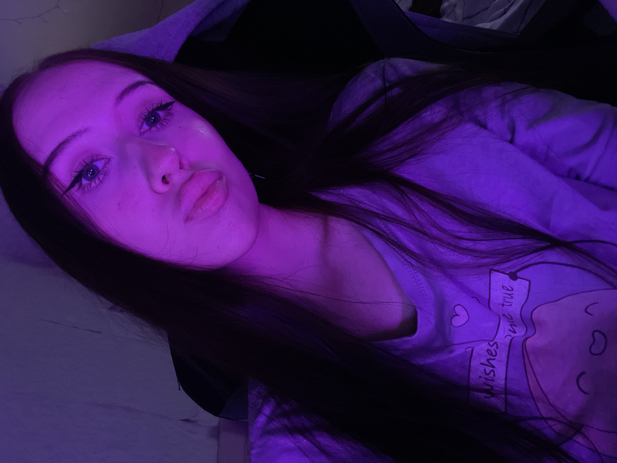 In an indoor setting with purple lighting, a young person with long dark hair is taking a selfie. They are wearing a light-colored t-shirt with a cartoon graphic and text that reads "wishes come true." Their face is angled towards the camera, with their lips pursed and their eyes looking towards the viewer. Eyeliner is visible on their eyelids. The background appears to be a wall and possibly furniture. There is no information in the image that would suggest a specific location in Votkinsk, Russia.