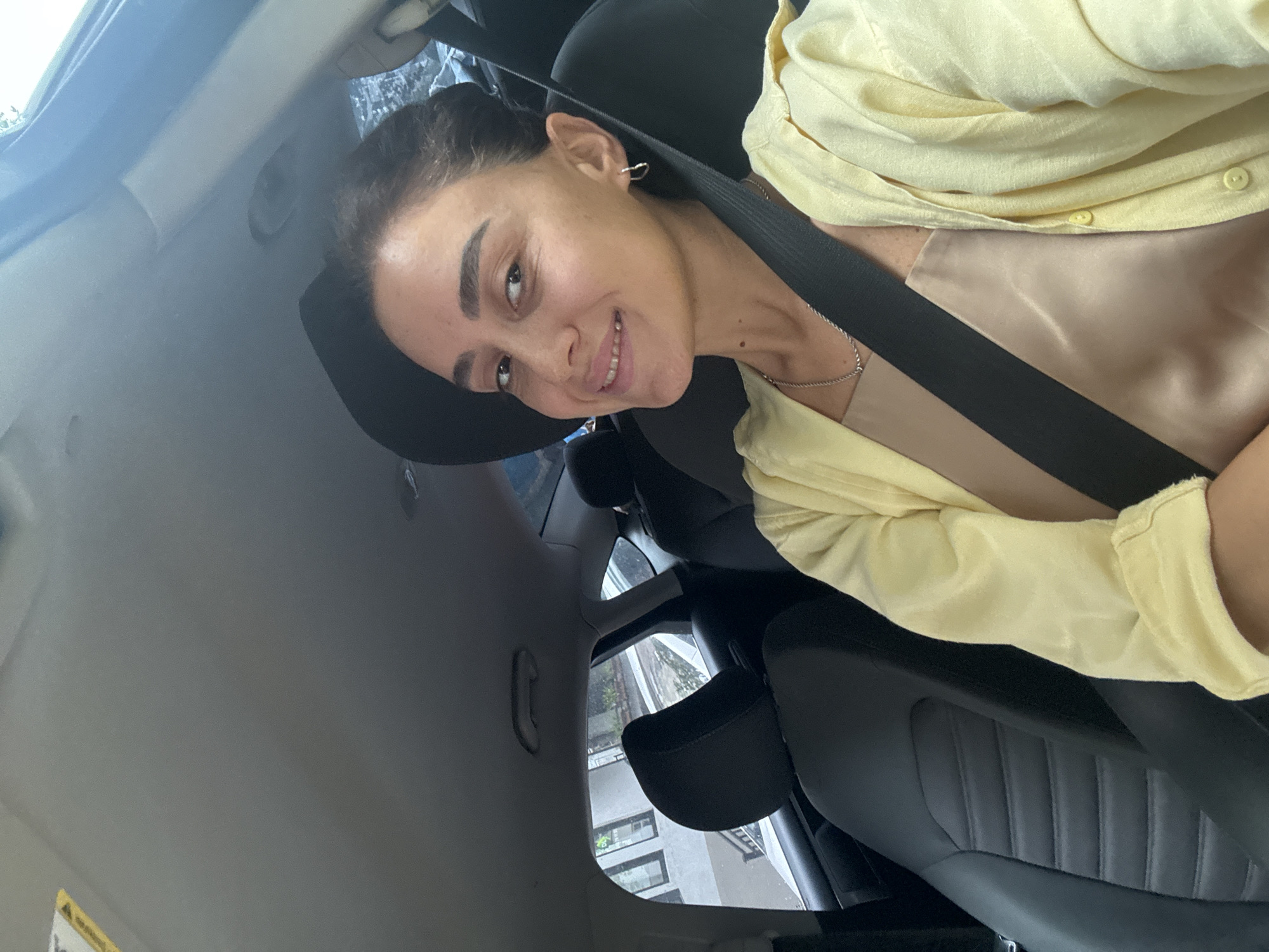 A woman is seen from the chest up, smiling and looking at the camera from the driver's seat of a car. She is wearing a yellow button-down shirt over a beige satin camisole and a silver necklace. A black seatbelt is visible across her chest. The interior of the car, including the dashboard and passenger seat, is visible. Outside the car window, buildings and trees are visible, suggesting an urban environment. The lighting indicates it is daytime.
