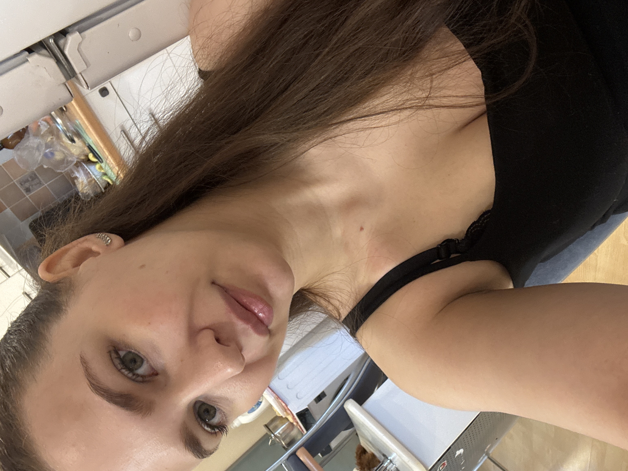 This image is a sideways-oriented selfie featuring a young woman captured from the chest up, in an indoor setting. The woman has long, straight, dark brown hair, fair skin, and light-colored eyes, appearing to be green or hazel. She has a neutral to gently smiling expression, looking directly into the camera with lightly glossed lips. She is wearing a black top, possibly a tank top or camisole, with a visible lace trim along the strap. A small, shiny earring or cuff adorns her upper left ear. A minor reddish mark or mole is present on her upper chest/collarbone area.
The background is a soft-focus interior space, revealing elements such as a light-colored shelf or cabinet, various small items stored in clear plastic containers, a section of small, light-colored tiles, and what appears to be part of a desk or workbench. A light switch or wall panel is also vaguely discernible. The setting suggests a casual, possibly domestic or practical workspace environment.
The primary activity is the woman taking her own photograph. The lighting is consistent with indoor ambient light, and no specific time of day can be determined. There is no legible text visible in the image. While the location context provided is Chaikovsky, Russia, the image itself does not contain specific visual cues to confirm this.