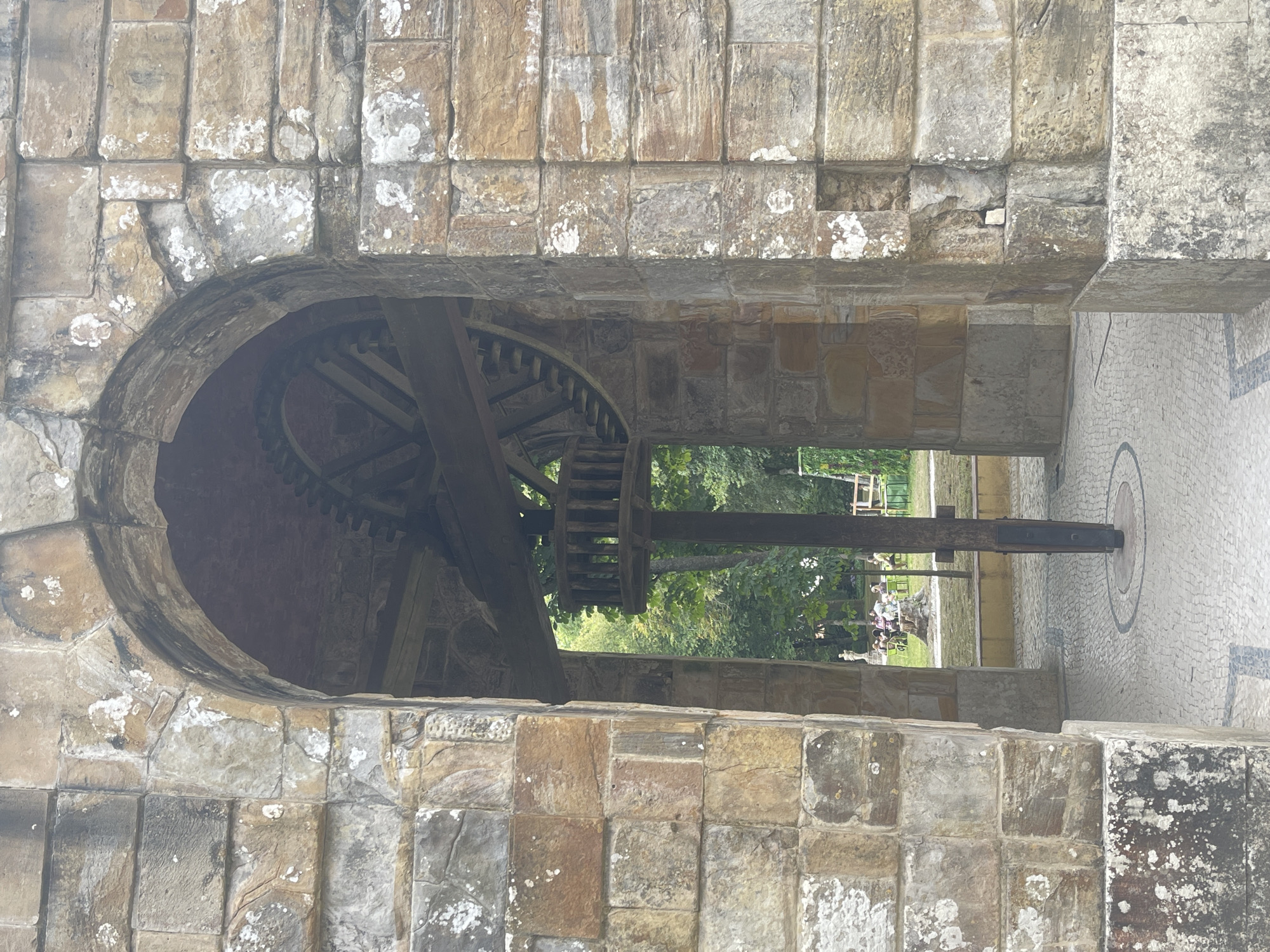 The image shows an arched stone gateway with an ancient wooden mechanism visible inside. The gateway is constructed of rough-hewn stone blocks of varying sizes and colors, ranging from light brown to grey, with patches of lichen. The archway frames a view of a park-like setting with green trees in the background.
Inside the arch, a large wooden gear is mounted on a horizontal beam. Below the gear, a vertical wooden post extends down to the ground, supporting a small circular wooden platform with vertical slats. The ground within the archway and extending outside is paved with small cobblestones arranged in a circular pattern around the base of the wooden post. In the background, through the arch, people are seated at tables under trees, suggesting a public space or cafe.
This scene is located at the **Tapada Nacional de Mafra** in Mafra, Portugal. This specific area is likely near the **Palace of Mafra**, within the grounds of the national hunting reserve. The wooden mechanism appears to be part of a historical water pump or other machinery associated with the estate's past. The overall impression is one of historical preservation within a natural setting.