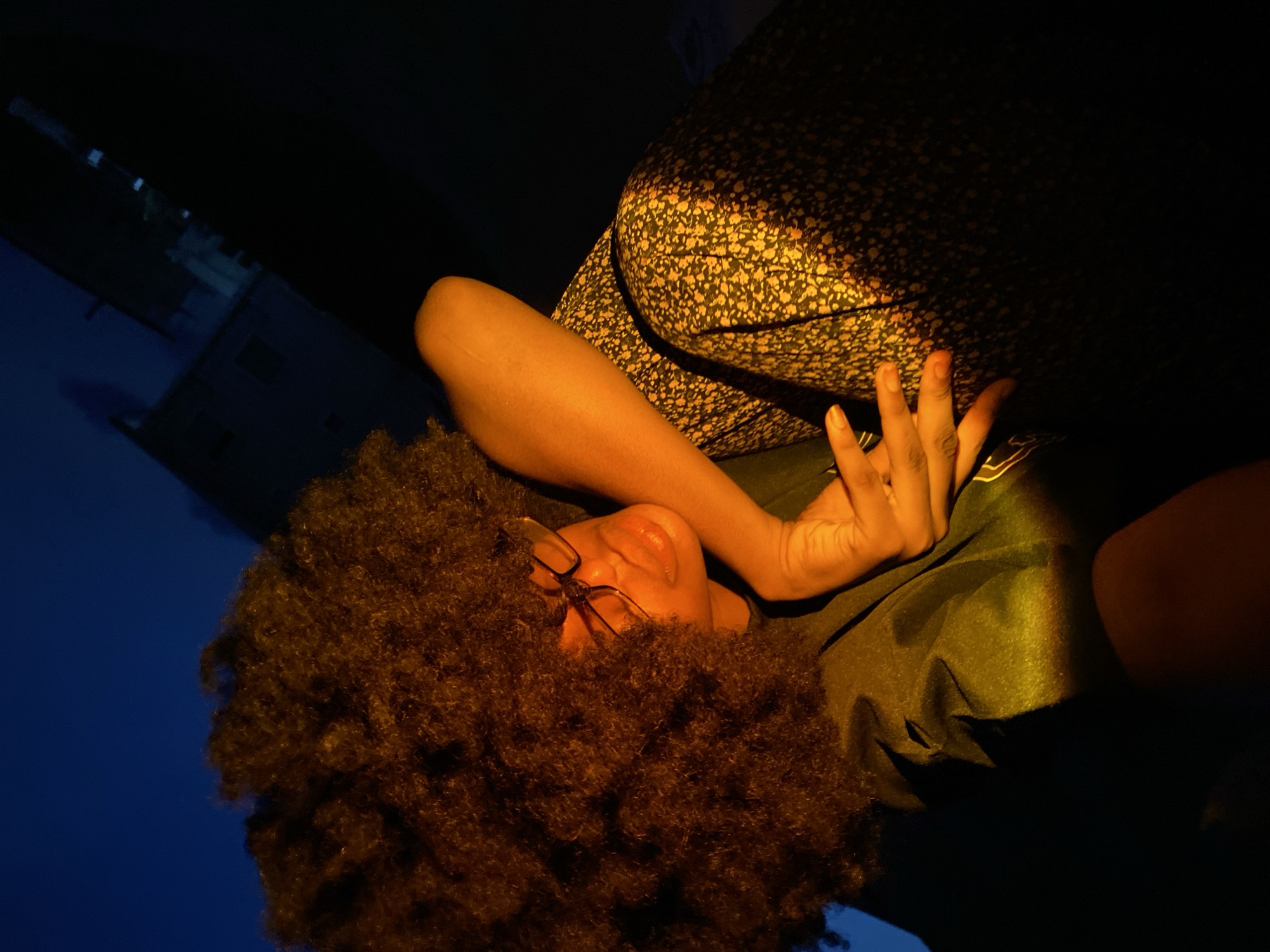 A single individual is depicted at night in Iranla, Nigeria. The subject, positioned off-center and angled, has dark skin, an expansive natural afro hairstyle, and wears eyeglasses. They are dressed in a dark top and a dark skirt patterned with small, light-colored specks. The person's left forearm and hand rest on their upper left thigh or knee. A strong, directional warm light source, originating from the lower right of the frame, illuminates the subject's face, hair, left arm, and the patterned skirt, creating high contrast with the surrounding darkness. The background shows a deep blue-black sky or environment, with faint, dimly lit structures resembling buildings visible in the upper right, partially silhouetted against the dark conditions.
