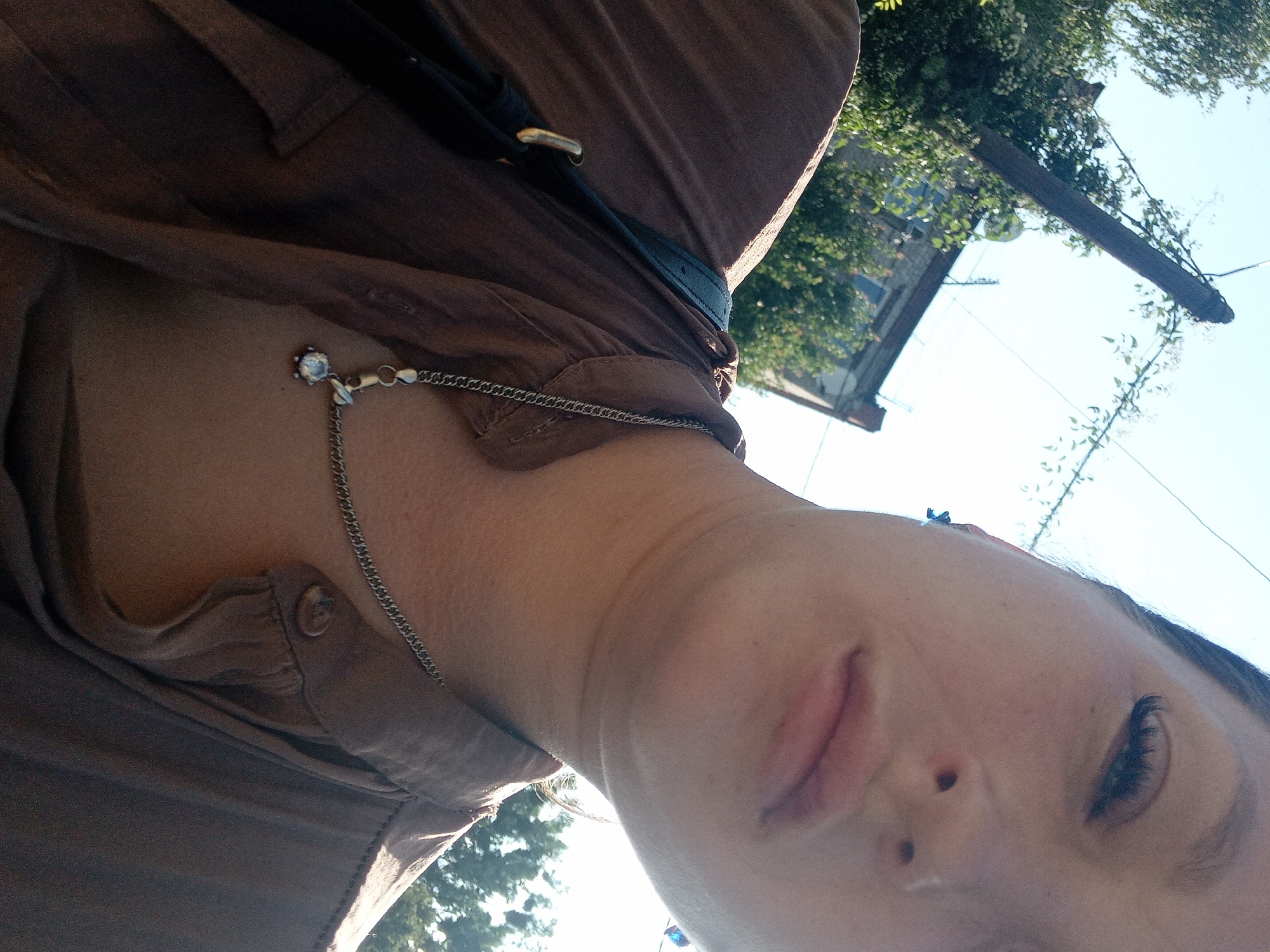 A close-up, low-angle outdoor photograph shows a woman's face and upper body. The woman is wearing a brown button-up shirt and a silver chain with a clear pendant. Her dark hair is pulled back. The background includes a clear blue sky, trees, and buildings, suggesting a residential area in Chuhuiv, Ukraine. A utility pole is visible in the upper left quadrant of the image. The lighting indicates it is daytime.