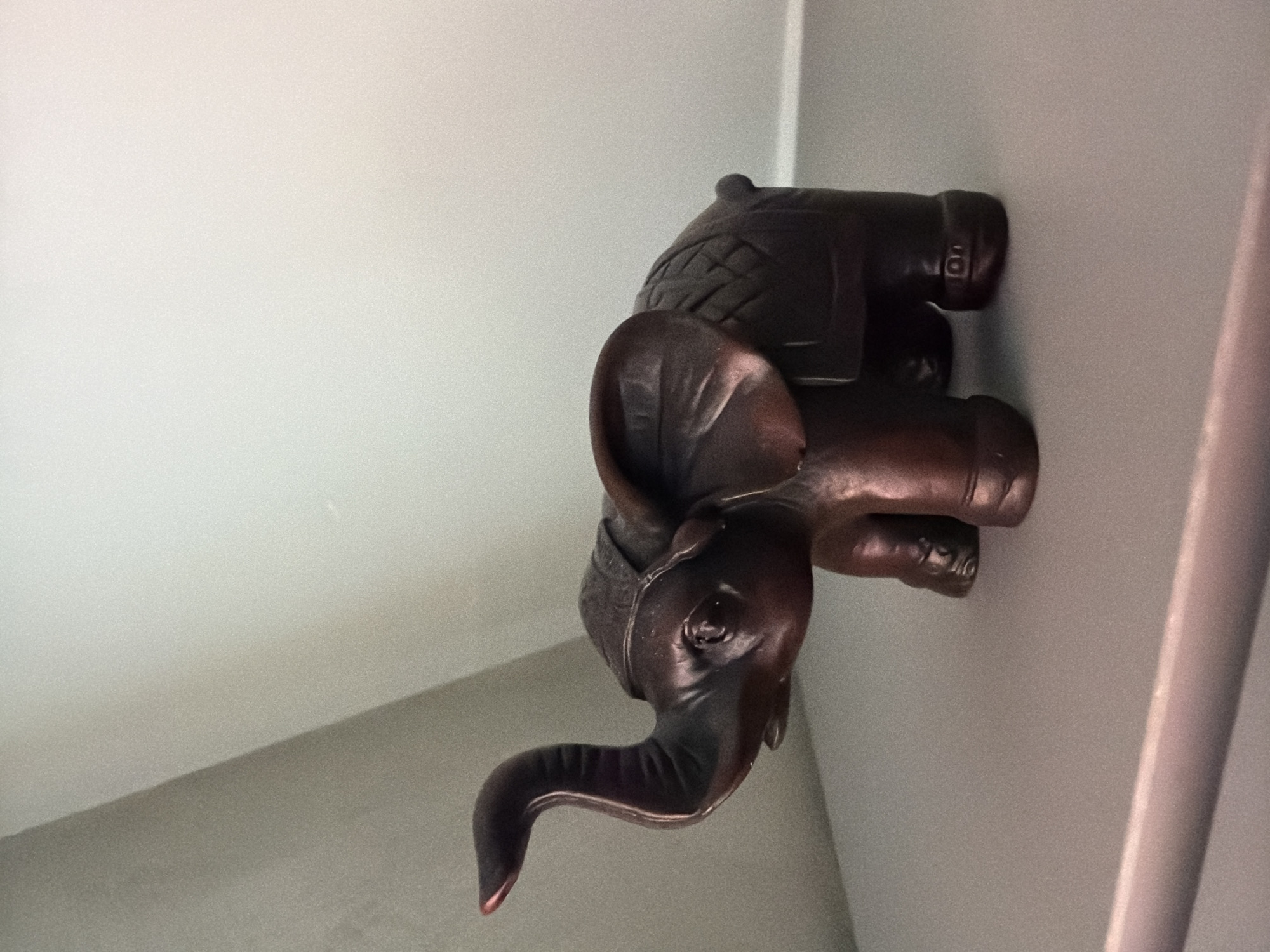 A dark-toned, possibly bronze or resin, elephant figurine is positioned on a light grey, matte surface within an interior setting in Sofia, Bulgaria. The figurine, depicted with its trunk raised and curled upwards, faces slightly towards the left. Distinct features include a decorative, textured saddle or blanket on its back, prominent ears, and visible eye details, with a subtle reddish hue on the tip of the trunk. The background comprises a corner formed by two walls: the left wall is a medium grey, while the right wall is a lighter, off-white color. Both wall surfaces and the foreground surface exhibit a fine granular texture. A thin, horizontal linear element is discernible in the lower foreground, indicating an edge or seam. Lighting appears to emanate from the right, casting illumination across the lighter wall and creating subtle shadows on the left side of the figurine and the darker wall.